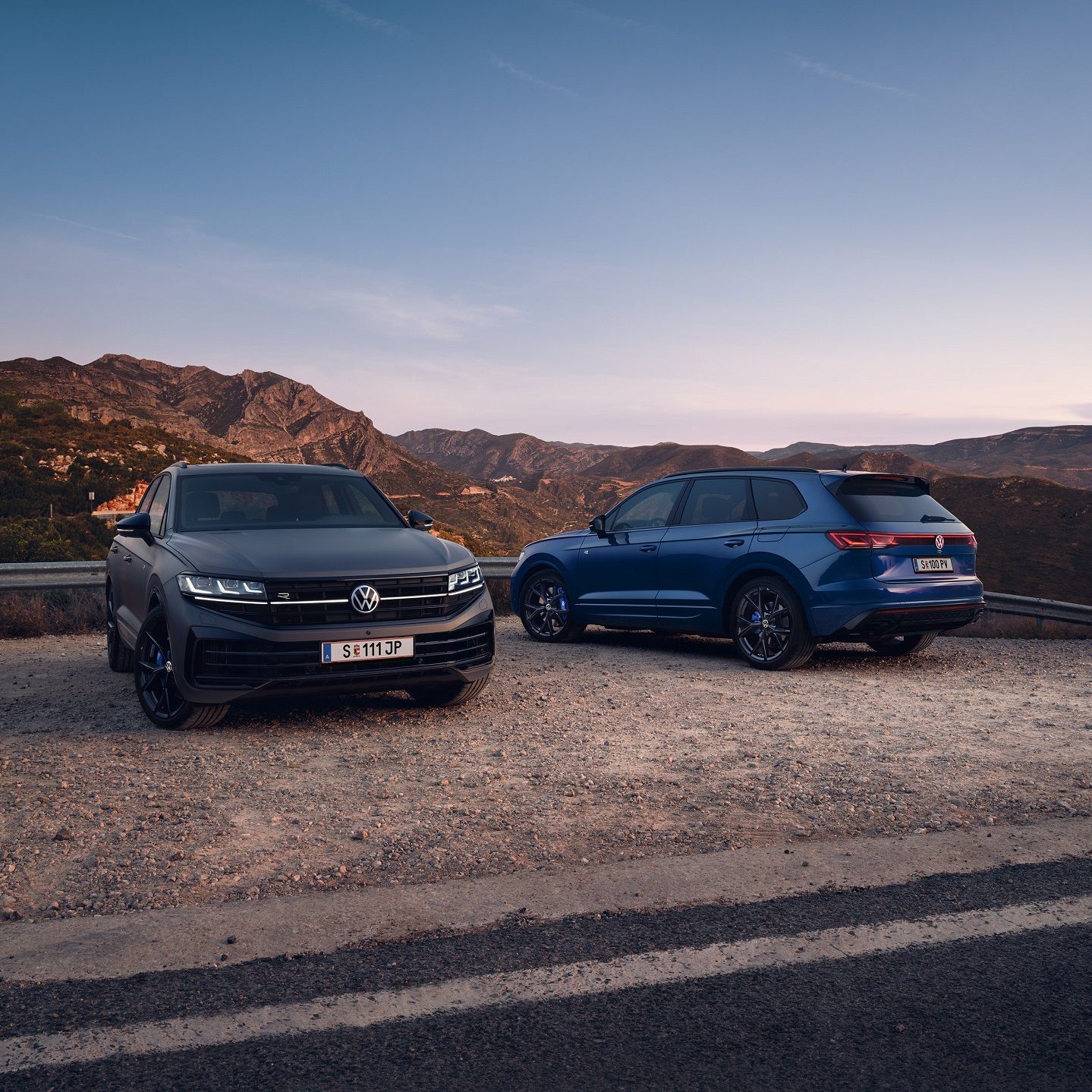 Dve vozidlá Volkswagen Touareg stoja na štrkovom priestranstve pri ceste. Z miesta je výhľad na pohorie.
