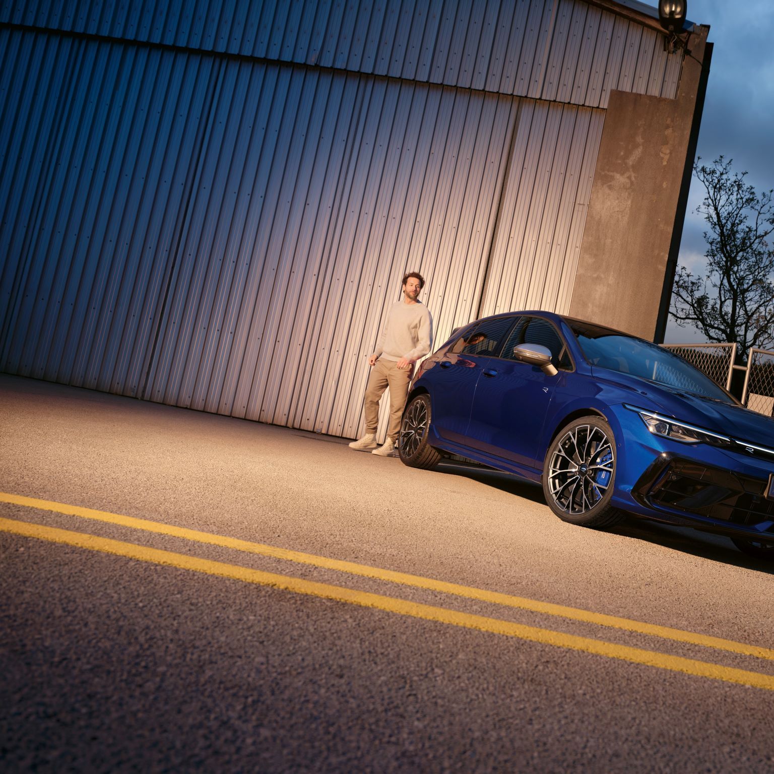 Modrý VW Golf R zaparkovaný pred industriálnou budovou a stojí pri ňom muž. Obloha je zatiahnutá, je šero a svieti pouličná lampa.
