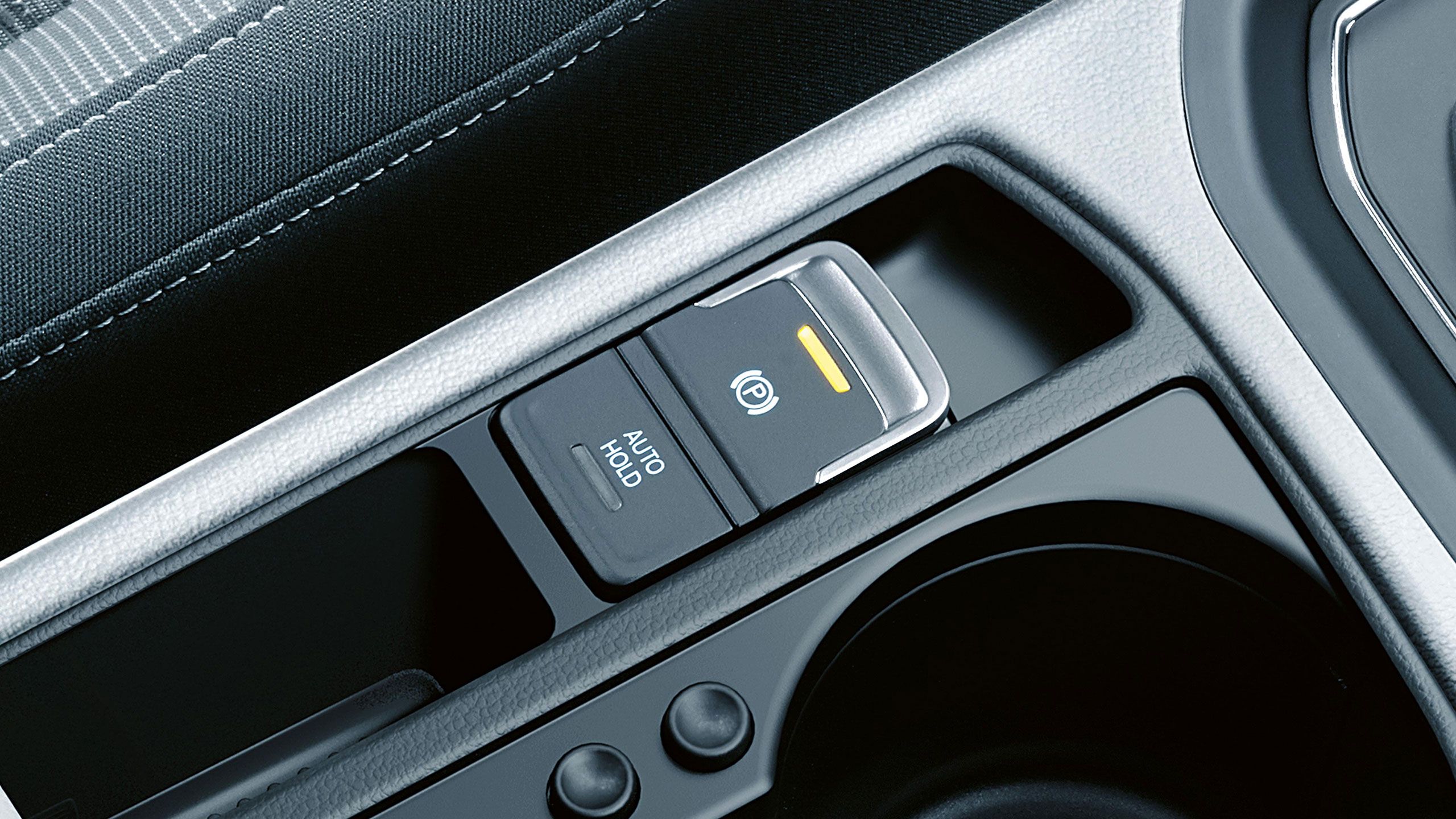 Detailný záber na ovládacie prvky v automobile, konkrétne tlačidlo elektronickej parkovacej brzdy a tlačidlo asistenta rozjadu do kopca "AUTO HOLD". Nachádzajú sa pri sebe v stredovej konzole.
