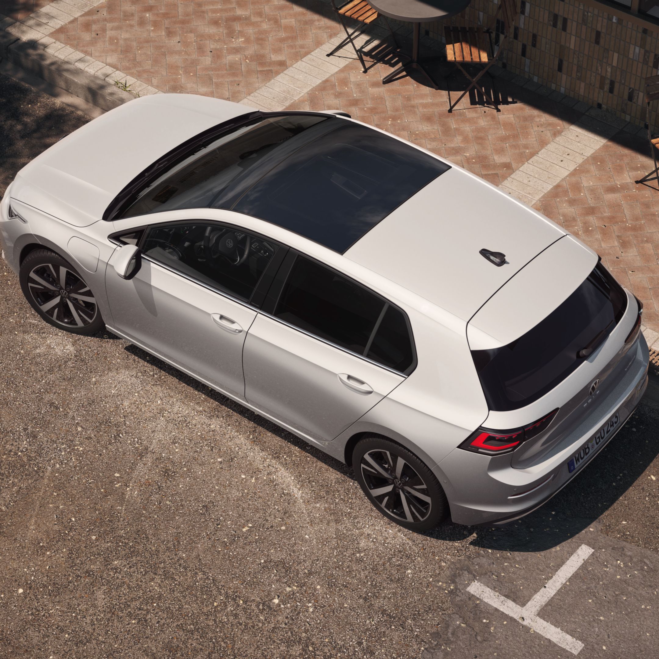 Pohľad zhora na biely VW Golf so zameraním na panoramatickú výklopnú/posuvnú strechu. Vozidlo stojí na parkovacom mieste pri chodníku v meste vedľa kaviarne.