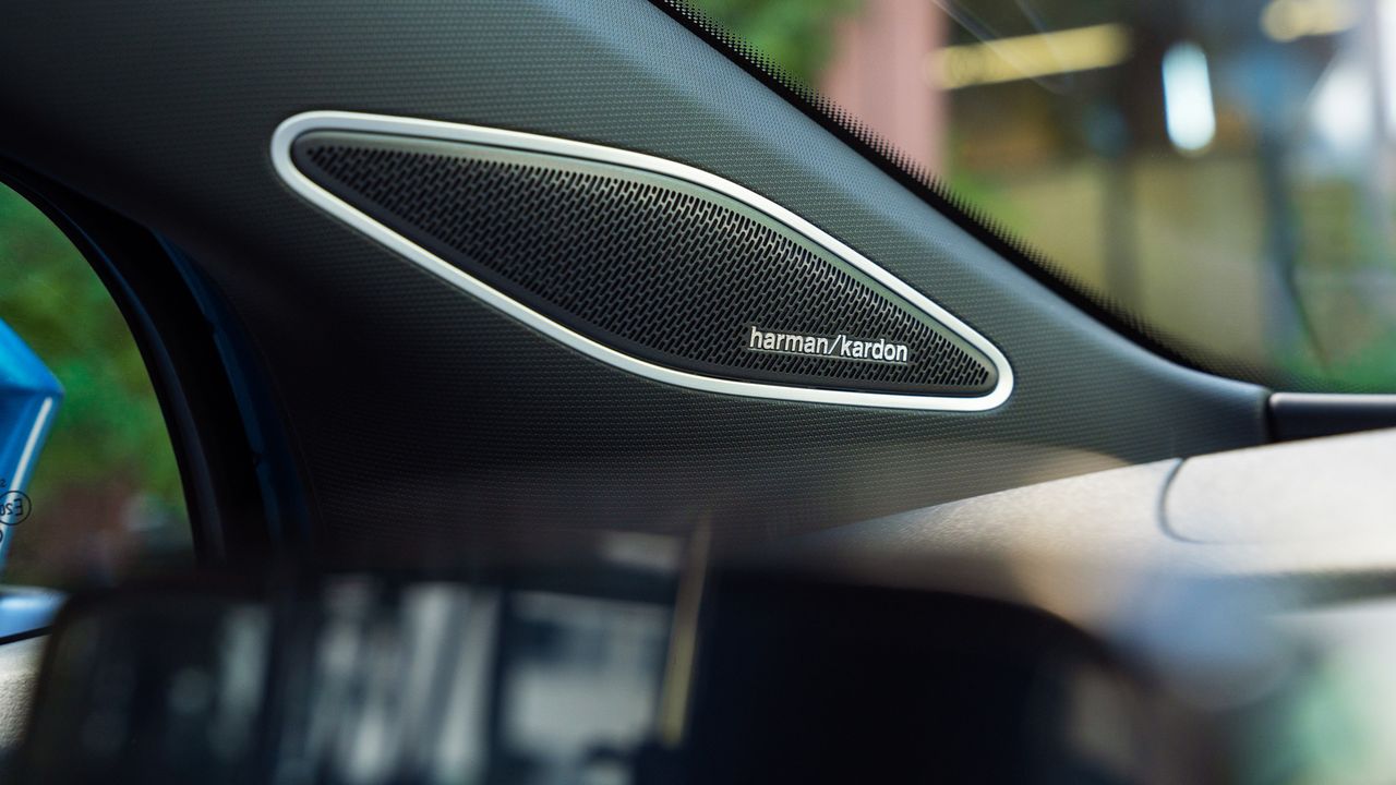 Pohľad na reproduktor Harman/Kardon integrovaný v A-stĺpiku. Mimo detailu je okno auta, cez ktoré čiastočne vidieť vonkajšie prostredie.