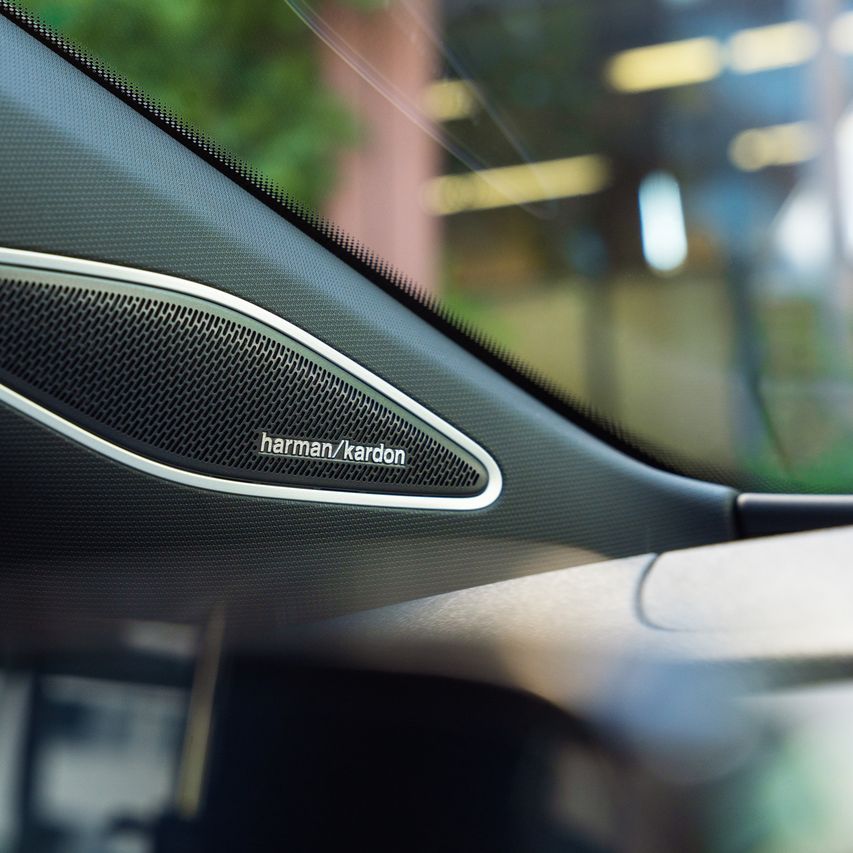 Pohľad na reproduktor Harman/Kardon integrovaný v A-stĺpiku. Mimo detailu je okno auta, cez ktoré čiastočne vidieť vonkajšie prostredie.