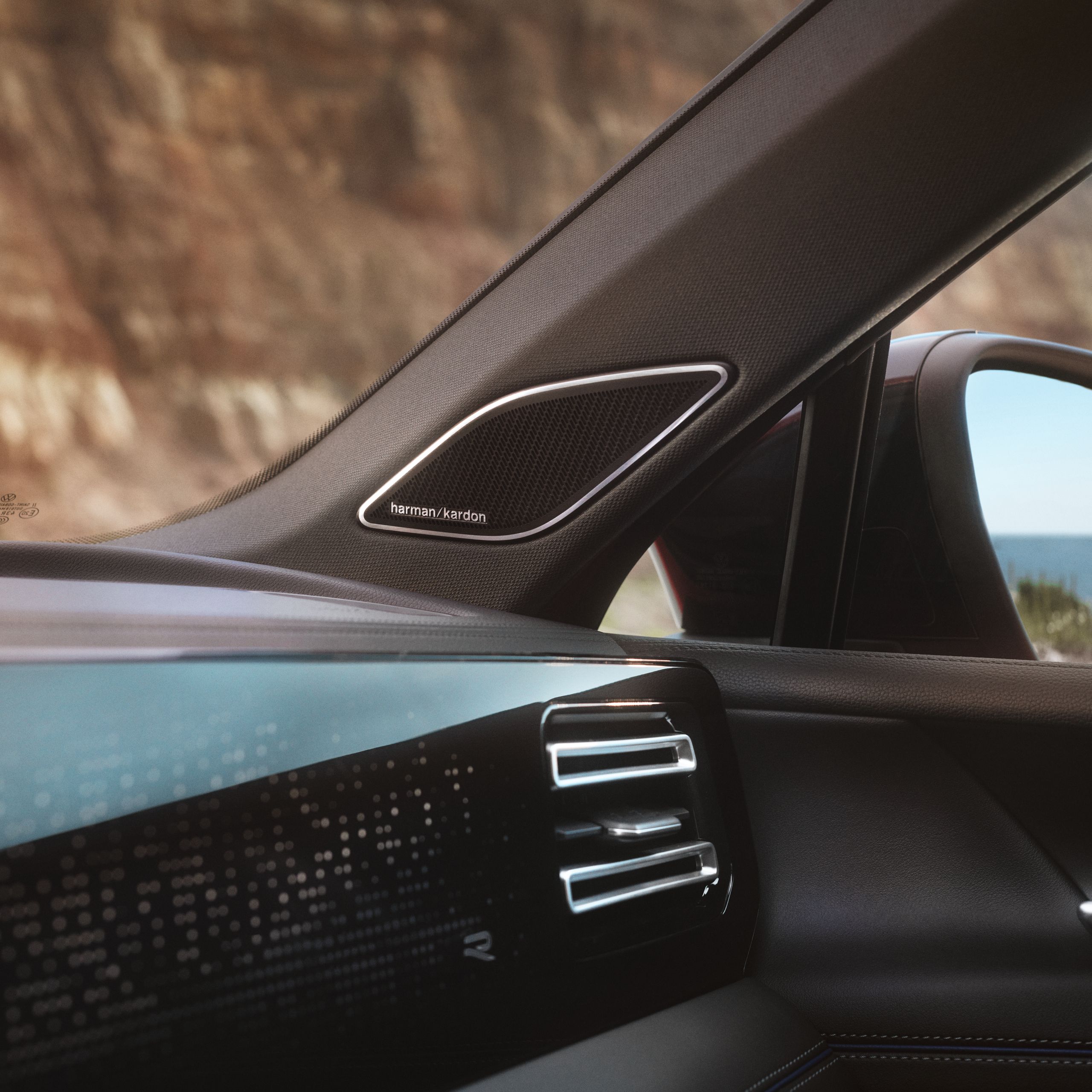 Detail interiéru VW Tiguan so zameraním na reproduktor inteegrovaný do A-stĺpika. Čiastočne vidieť palubnú dosku s ambientným ovládaním, vonkajšie spätné zrkadlo a cez okná výhľad na útes.