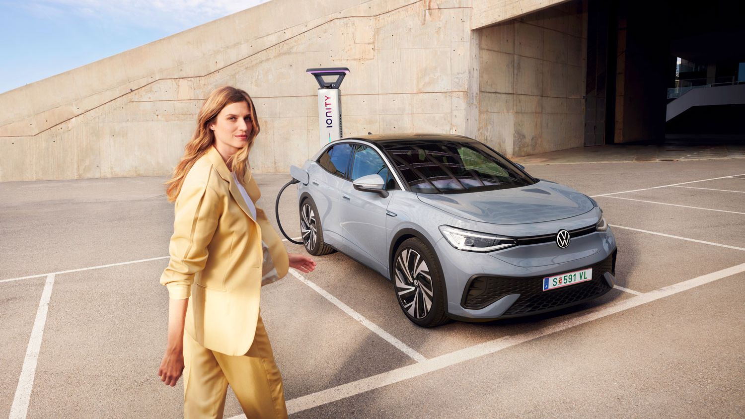 VW ID.5 v sivej farbe stojí na parkovisku a nabíja sa na nabíjacej stanici, pohľad spredu jemne zboku, žena v žltom kostýme kráča k vozidlu.