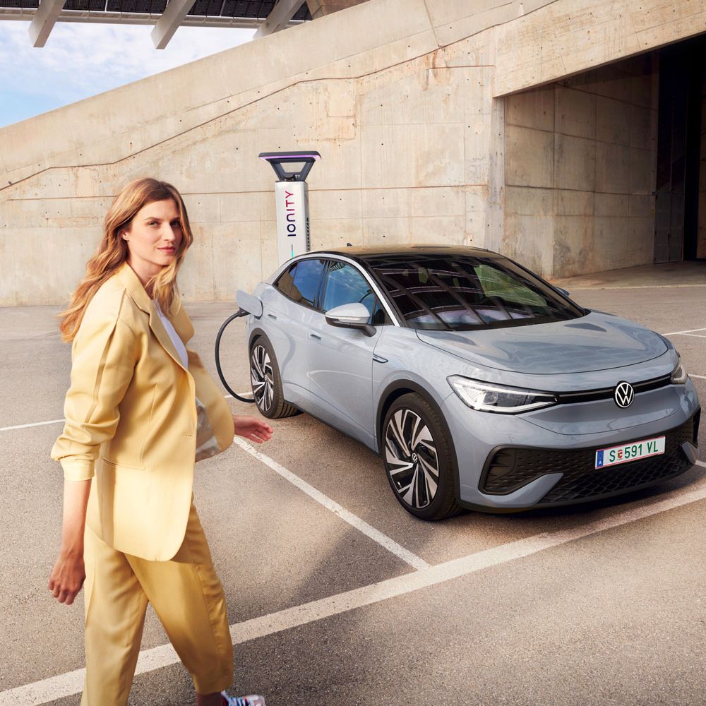 VW ID.5 v sivej farbe stojí na parkovisku a nabíja sa na nabíjacej stanici, pohľad spredu a zboku, žena kráča k vozidlu.