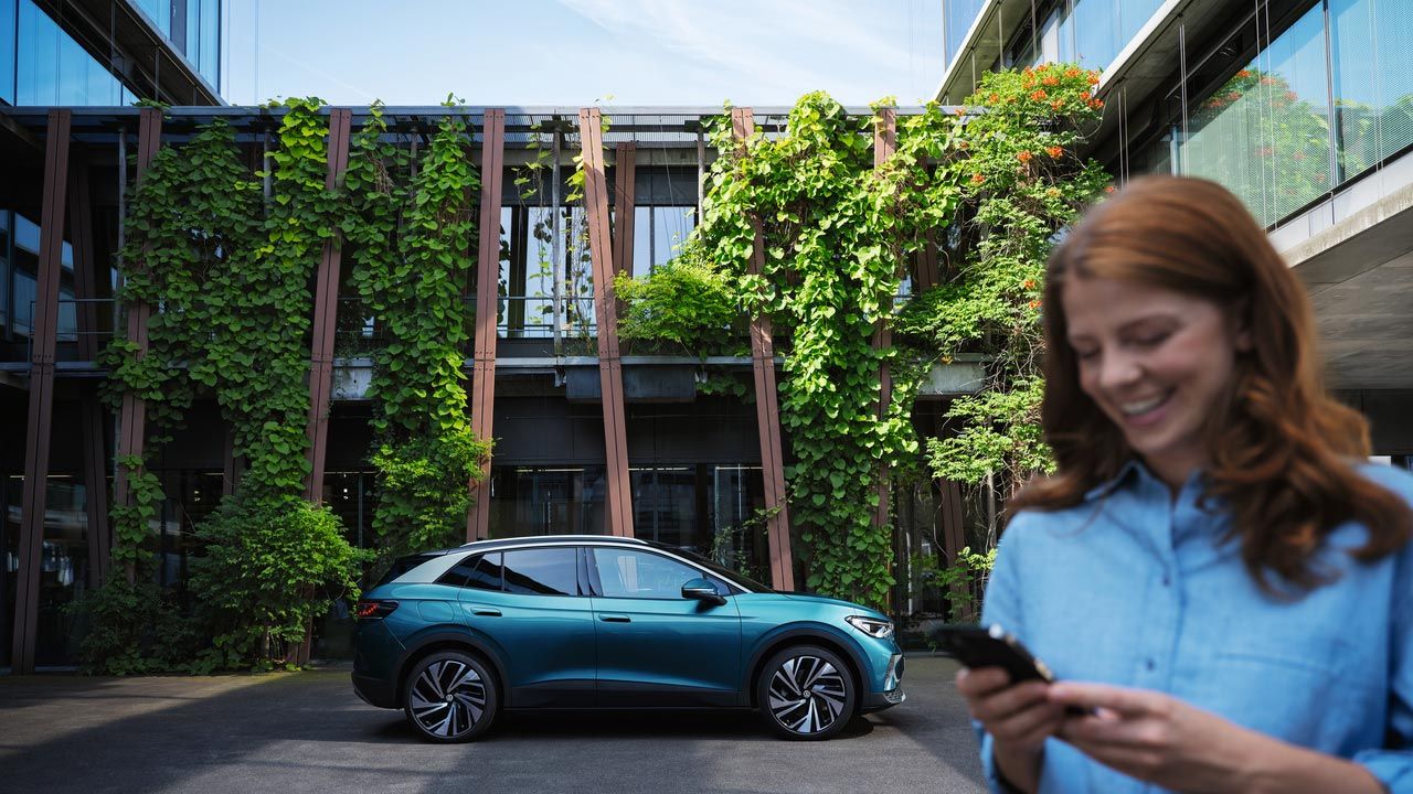 Mladá žena stojí na dvore pred vozidlom VW ID.4 Pro. V ruke drží mobilný telefón.