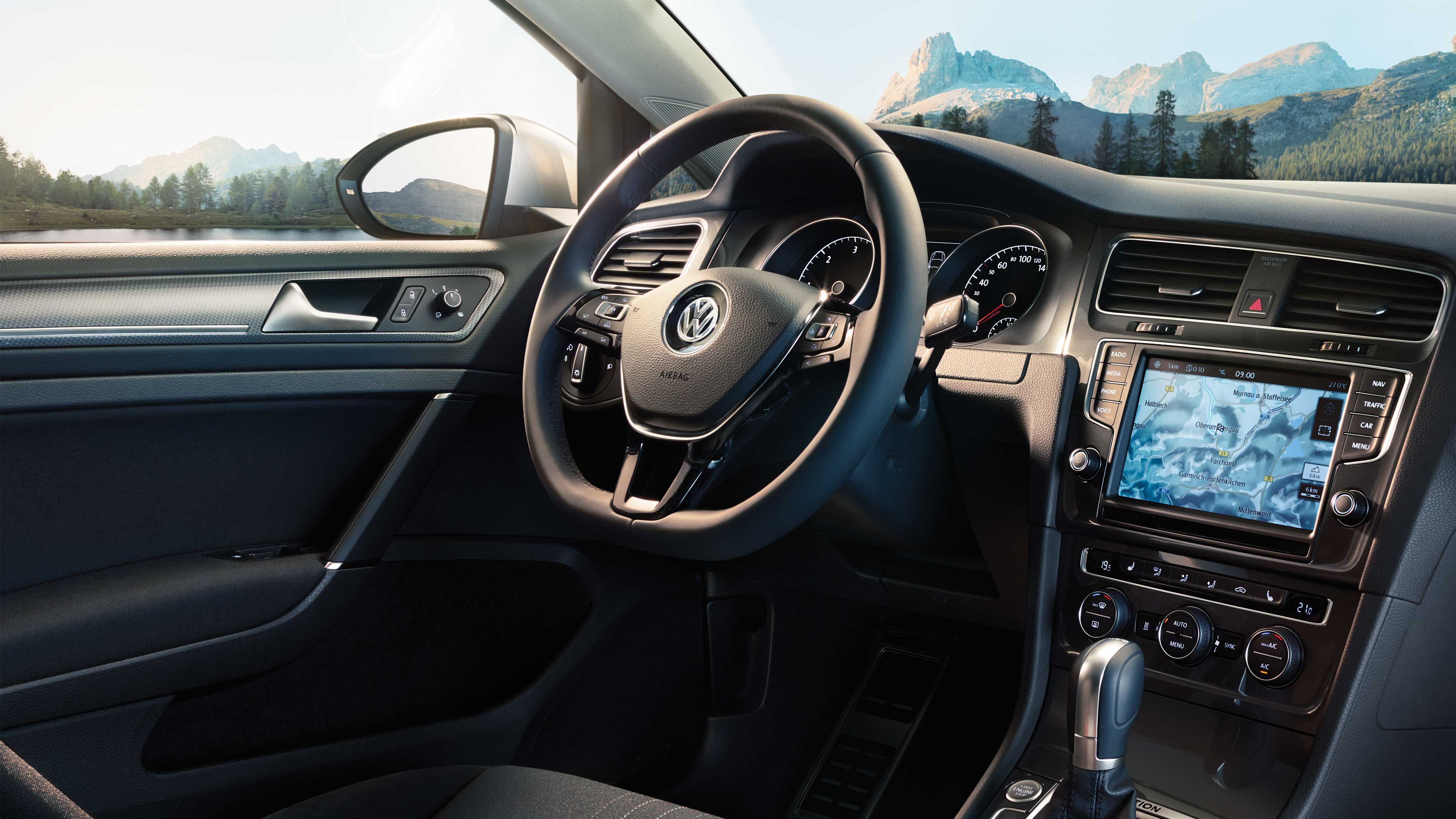 Interiér moderného automobilu Volkswagen so zobrazením volantu, palubnej dosky a navigačného systému na displeji v stredovej konzole. V pozadí vidieť scenériu hôr, lesa a jazera.