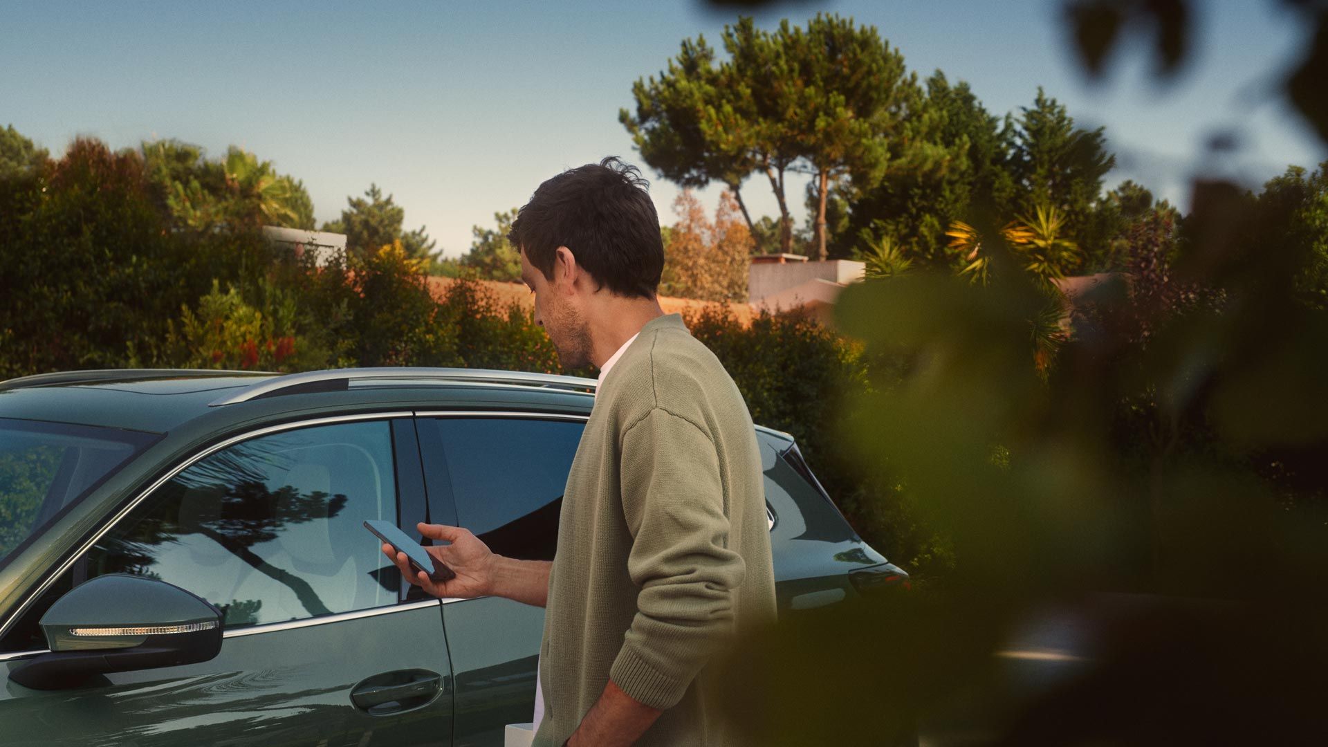 Mladý muž stojí na strane vodiča tmavozeleného VW Tiguan a pomocou smartfónu ovláda systém Park Assist Plus.