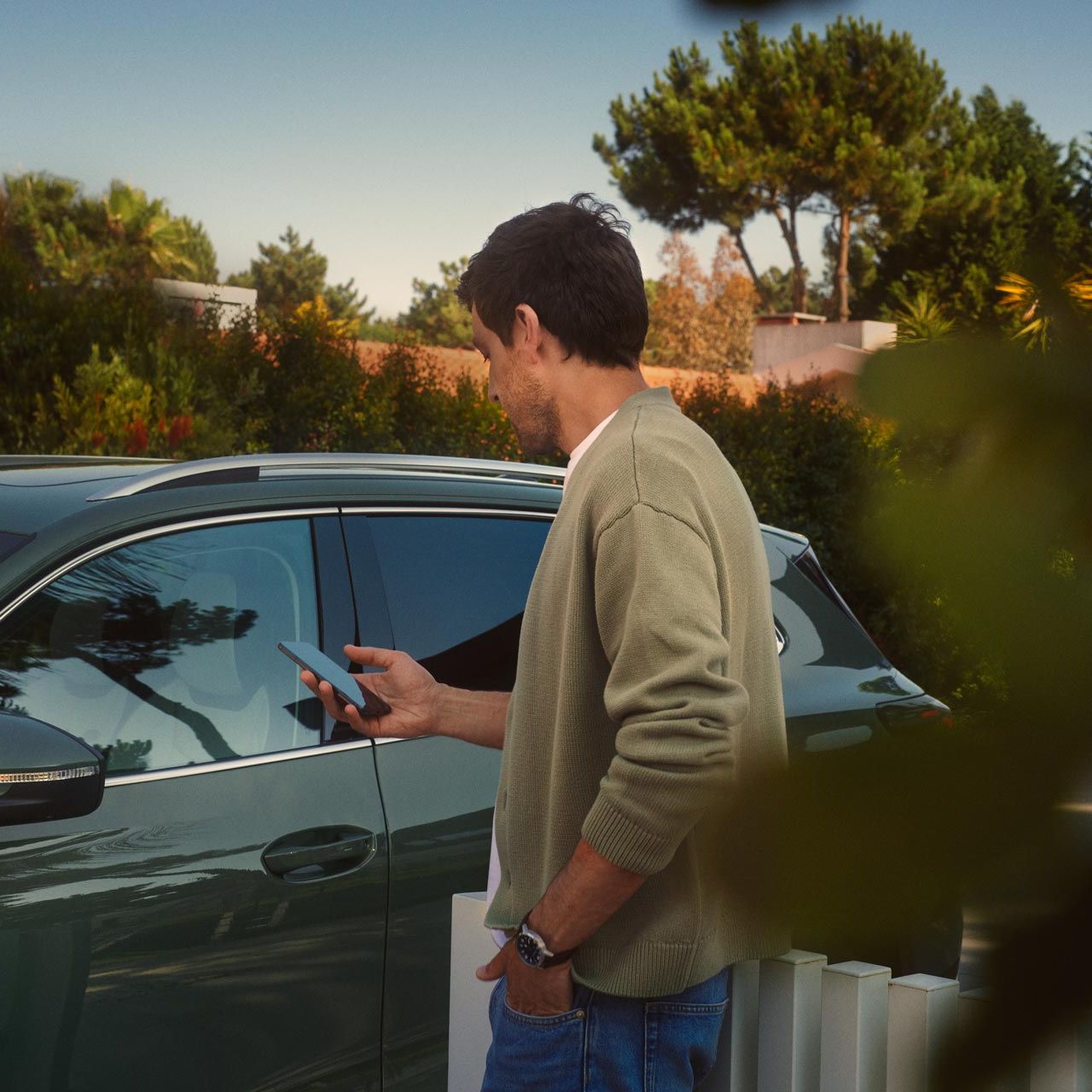 Mladý muž stojí na strane vodiča tmavozeleného VW Tiguan a pomocou smartfónu ovláda systém Park Assist Plus.