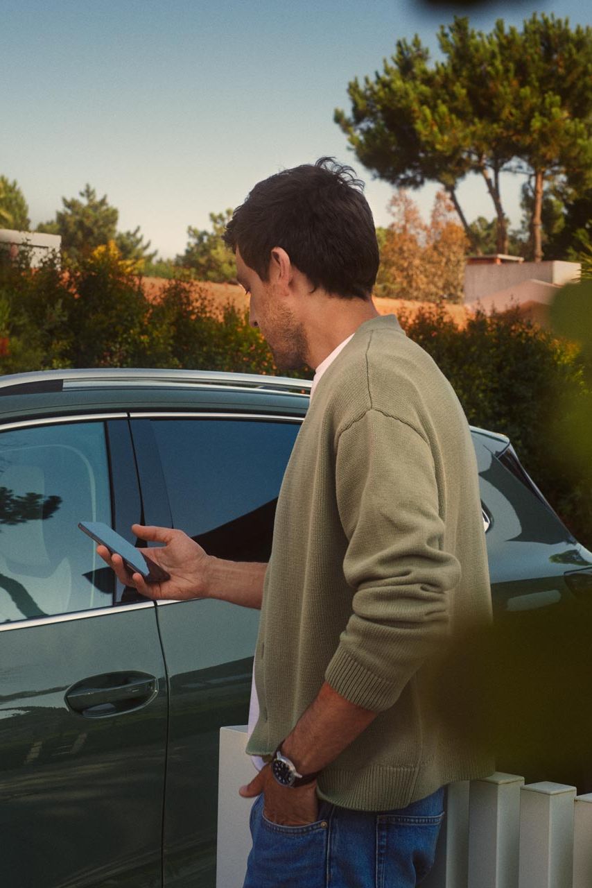 Mladý muž stojí na strane vodiča tmavozeleného VW Tiguan a pomocou smartfónu ovláda systém Park Assist Plus.
