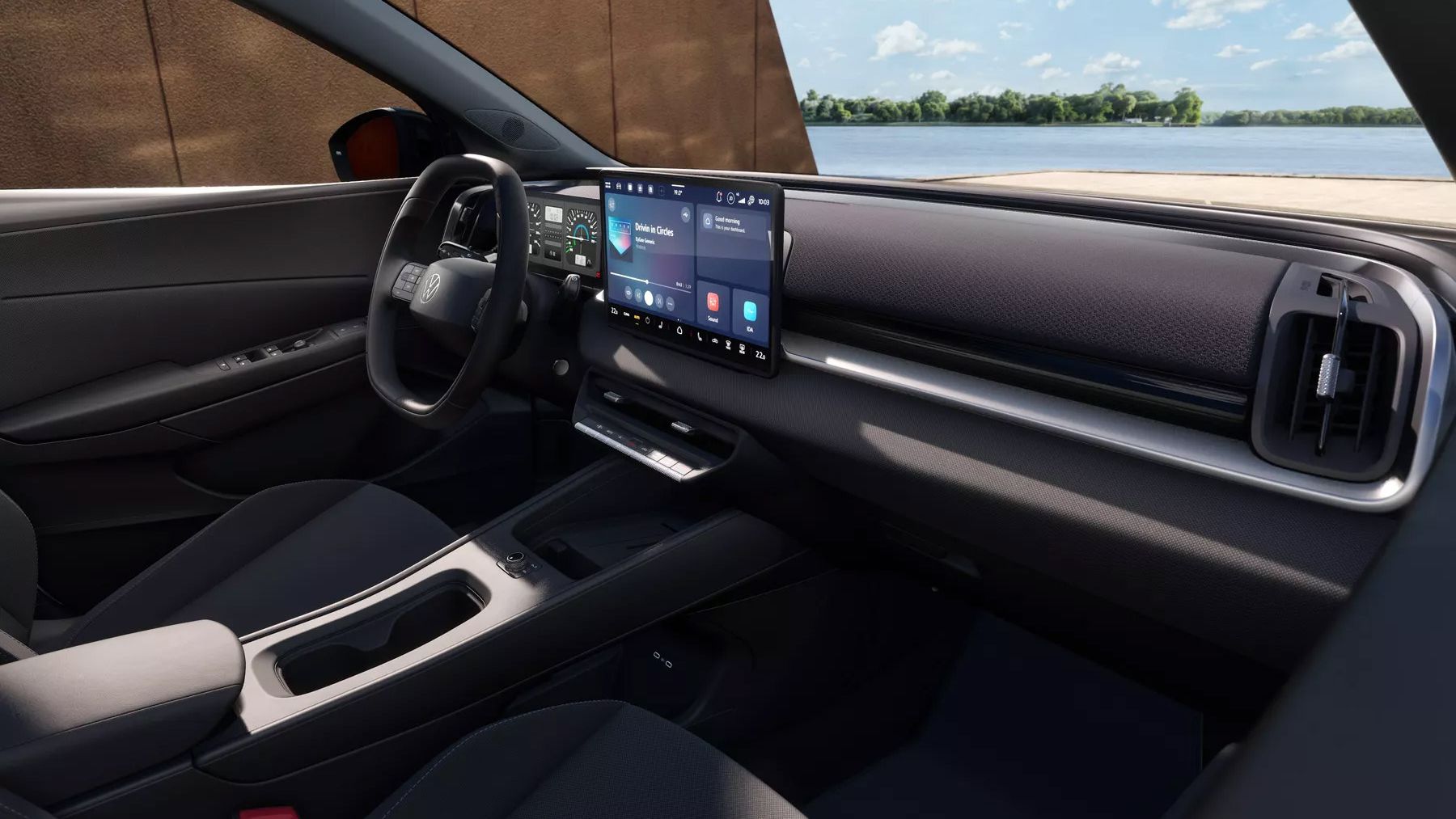 Moderný kokpit modelu VW ID. Cross Life s tmavým interiérom a veľkým dotykovým displejom pre infotainment umiestneným uprostred. Vľavo za volantom je vidieť digitálny prístrojový panel, vpravo jednoduchý palubný panel s integrovanými výduchmi vzduchu. Cez okná je vidieť slnečný deň s vodou a stromami v pozadí.