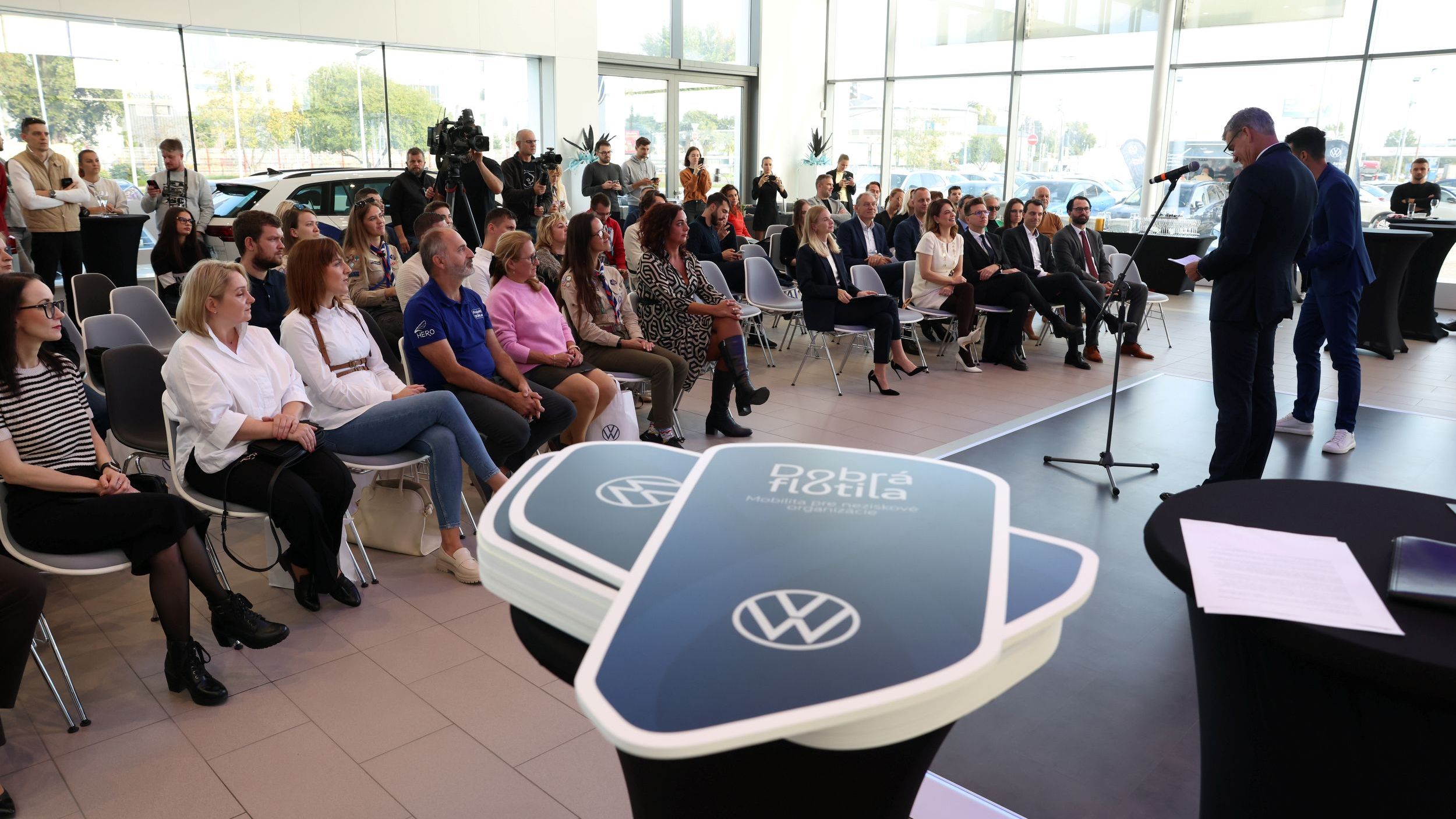 Skupina ľudí sedí na stoličkách v modernej, svetlej miestnosti, zatiaľ čo dvaja muži na pódiu prezentujú. V popredí sú umiestnené stojany s logom značky Volkswagen na banneroch s logom "Dobrá flotila".