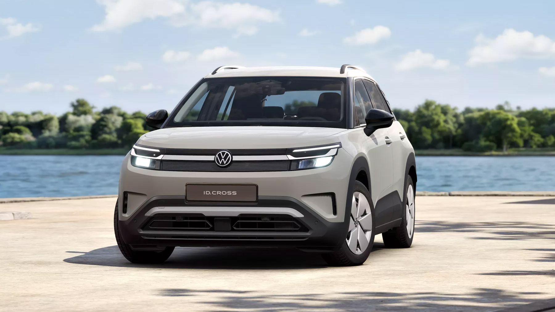 Svetlý, moderný VW ID. Cross stojí na nábrežnej promenáde pred jazerom, v pozadí sú stromy a modrá obloha s niekoľkými mrakmi. Auto je zobrazené z čelného pohľadu v miernom uhle, s LED prednými svetlometmi a logom VW uprostred.