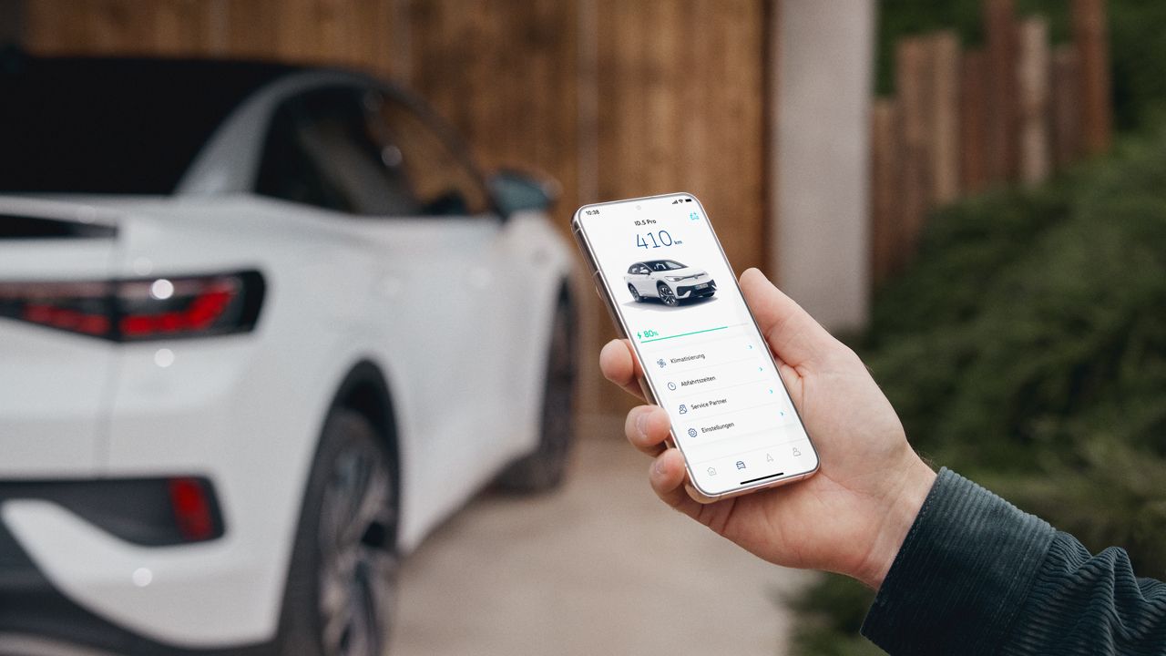 Muž drží smartfón, na ktorého displeji je zobrazená aplikácia na ovládanie elektrického vozidla. V pozadí je biele elektrické vozidla zaparkované pred drevenou bránou. Rovnaký automobil je zobrazený aj na displeji telefónu.