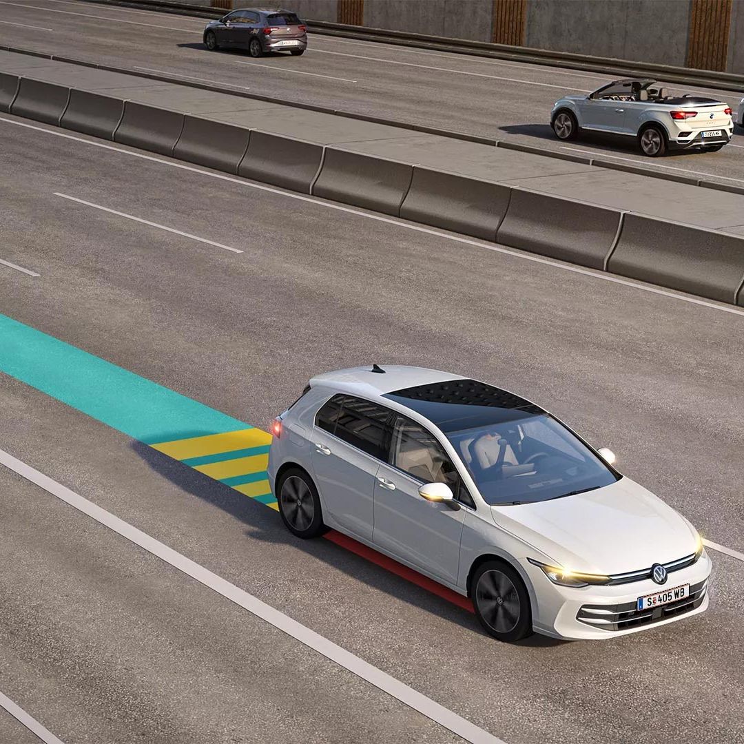 Biely VW Golf jazdí v premávke s aktívnym systémom Emergency Assist, schematicky znázornený na obrázku modro-žltým pásom.