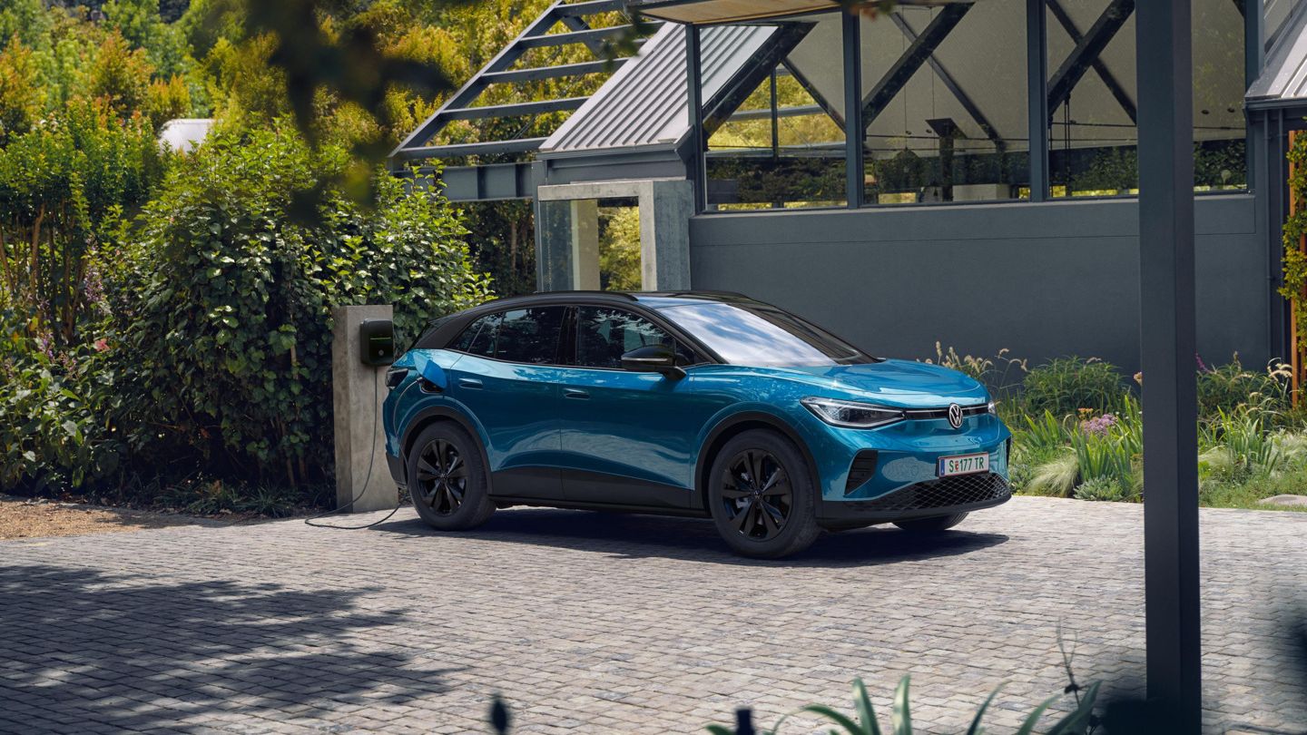 Modrý VW ID.4 s čiernou strechou parkuje vedľa moderného domu a nabíja sa na nabíjacej stanici. 