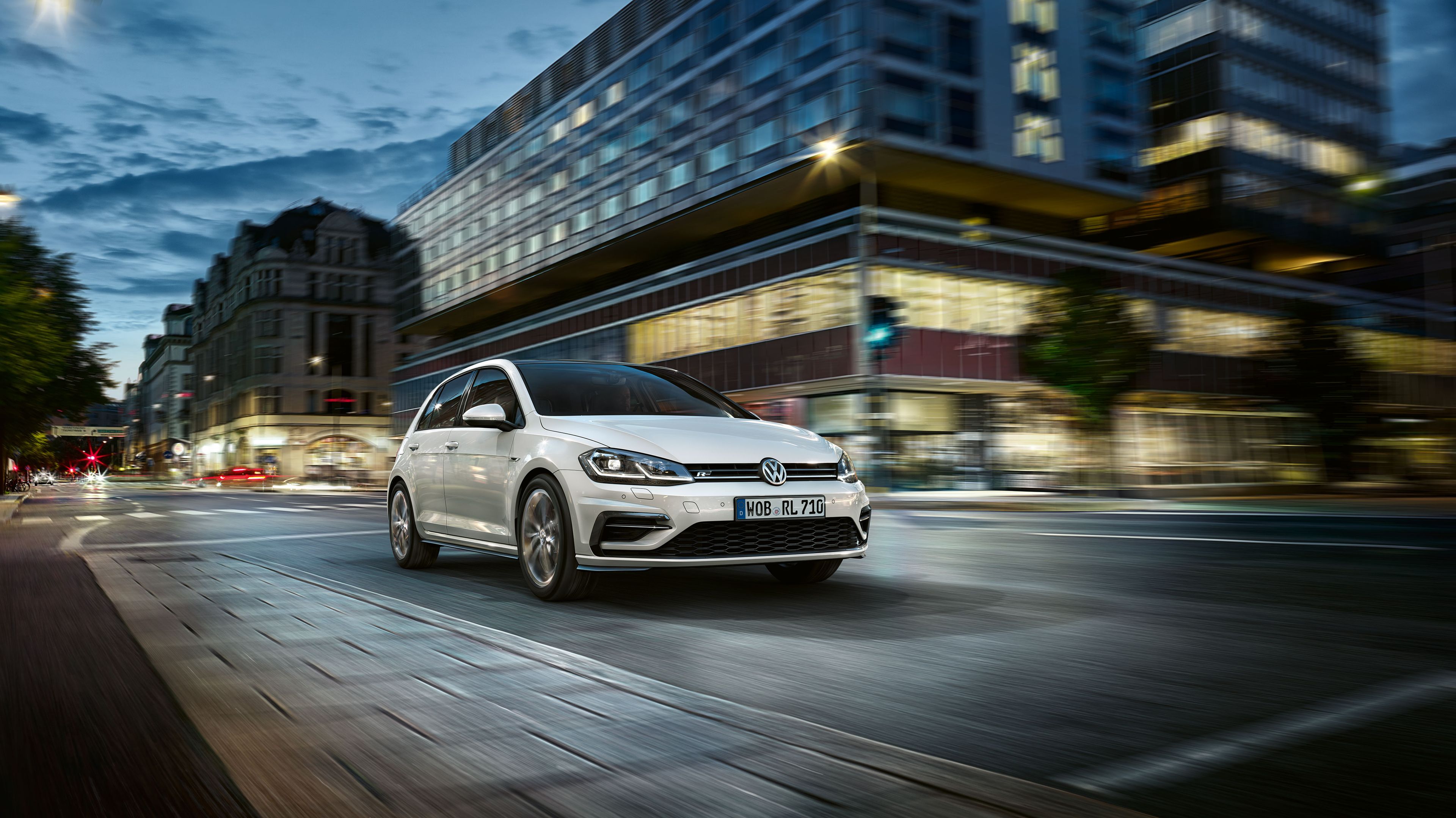 Biele vozidlo Volkswagen jazdí po modernej mestskej ulici počas večera. Budovy v pozadí sú osvetlené a svetlá na ulici vytvárajú dynamickú atmosféru. Auto je zachytené v pohybe.