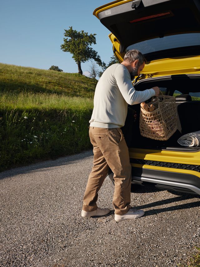 Muž vkladá veľký piknikový kôš do batožinového priestoru žltého VW T-Cross, ktorý je zaparkovaný na ceste v hornatej krajine.