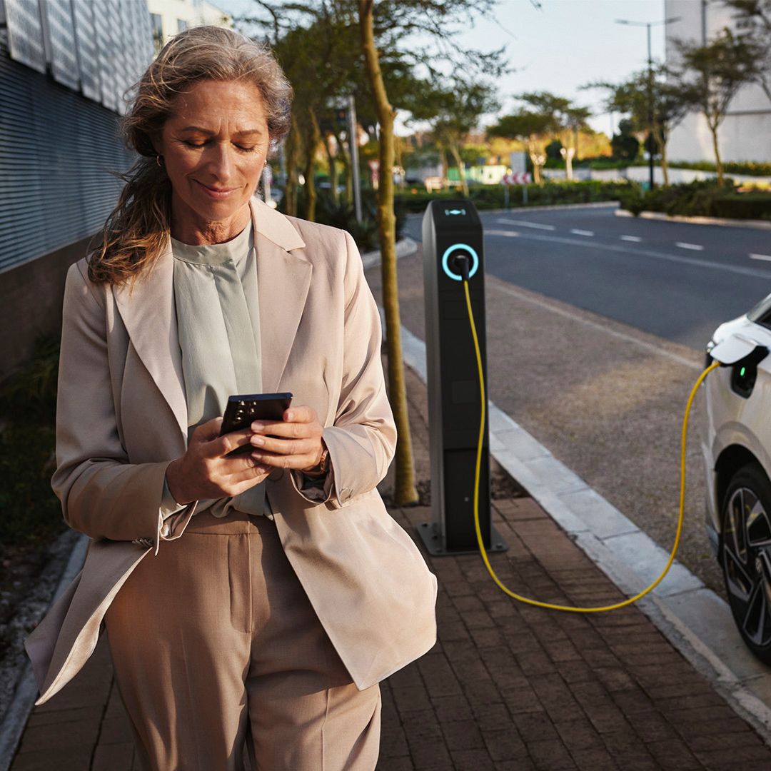 Žena v svetlom béžovom kostýme stojí pri vozidle, ktoré sa nabíja na nabíjacej stanici a drží mobilný telefón. Vozidlo je zaparkované pri chodníku. V pozadí je mestská scenéria moderného mesta.