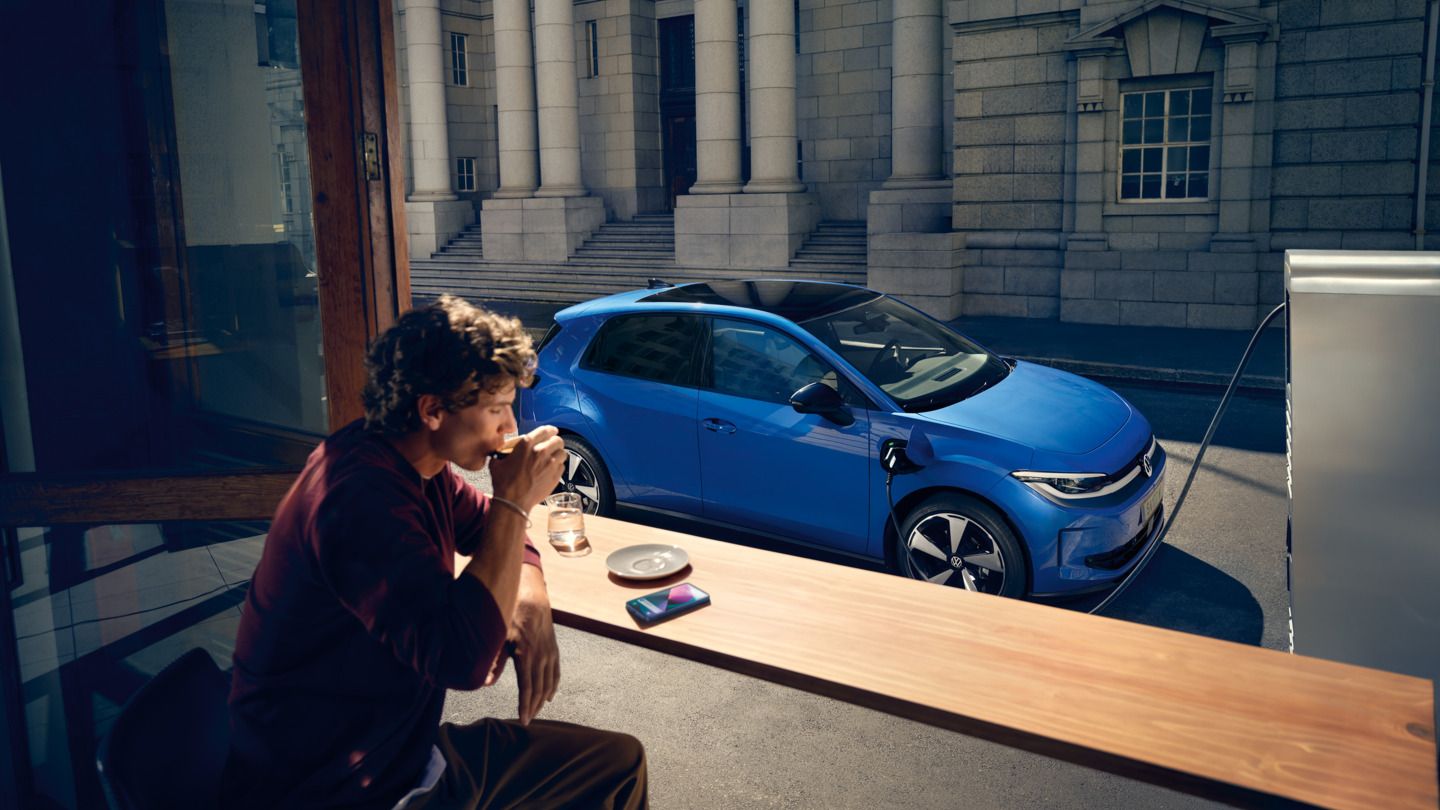 Muž sedí v kaviarni a cez otvorené okno je vidieť modrý VW ID. Polo Style, ktorý sa nabíja na nabíjacej stanici.