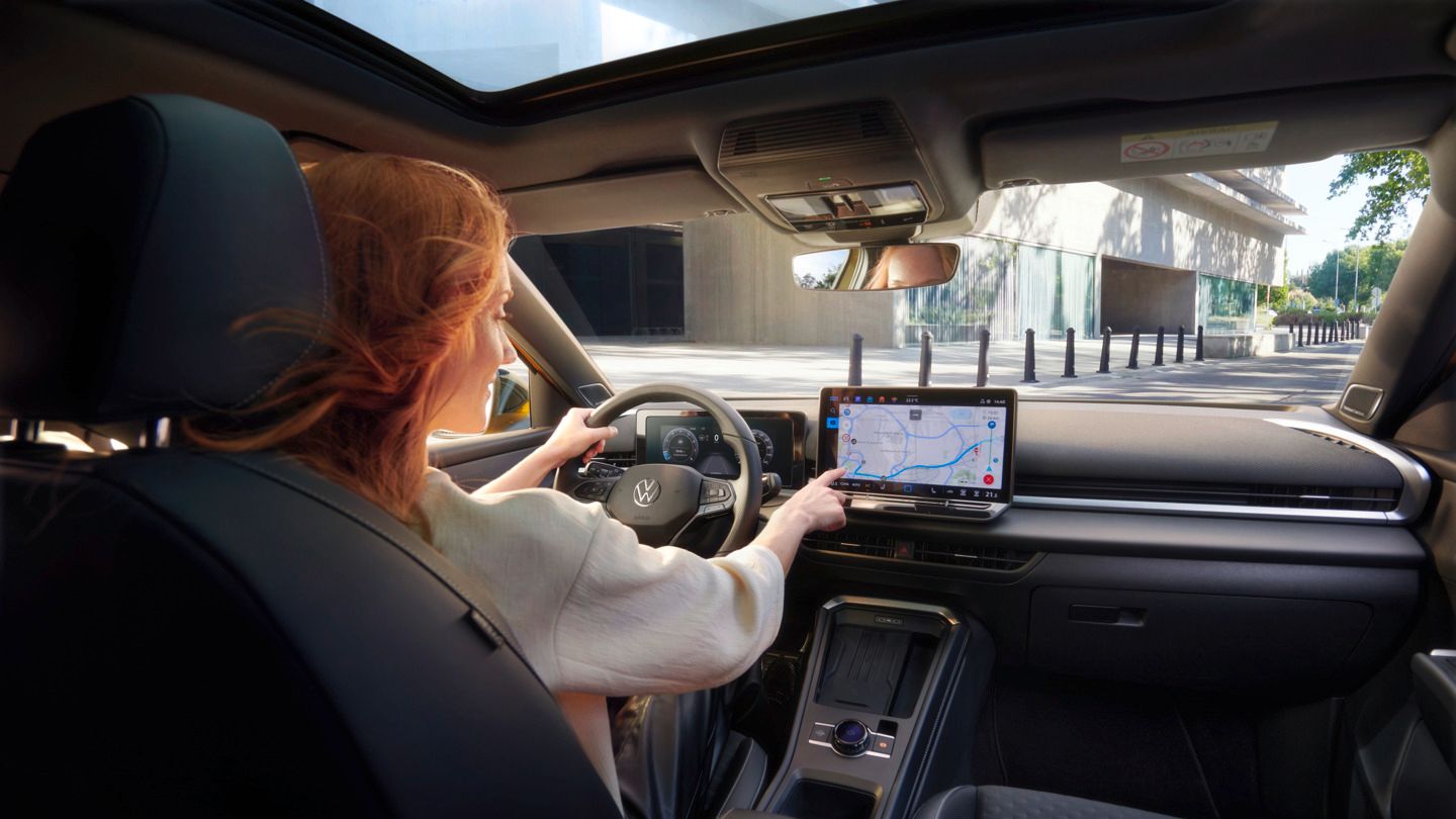 VW T-Roc Červenovlasá žena sedí na sedadle vodiča vo vozidle VW T-Roc. Vidieť prednú polovicu čierneho interiéru. Jednou rukou ovláda navigáciu v infotainment systéme prostredníctvom dotykovej obrazovky, druhou rukou drží volant. Vozidlo sa zdá byť v meste.