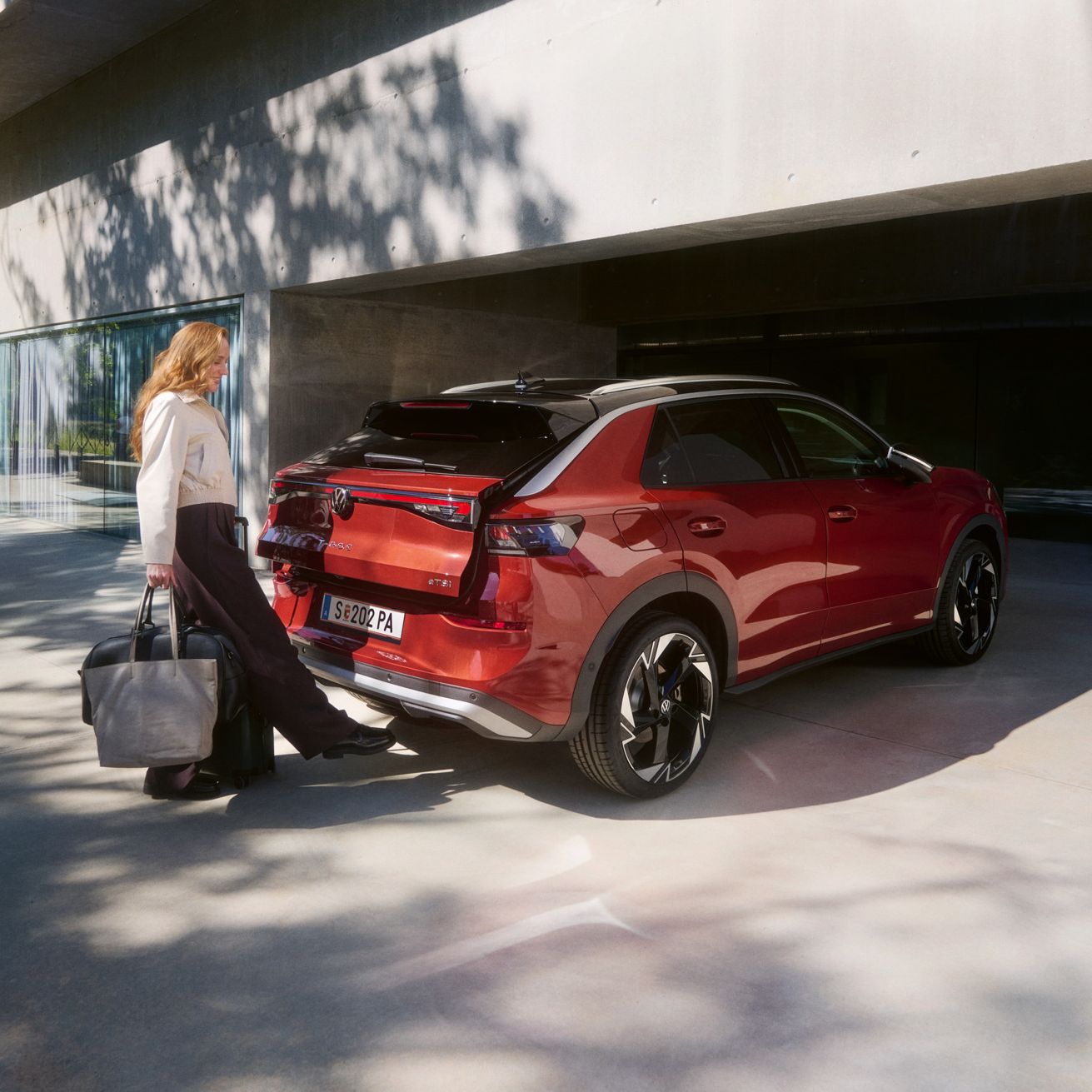 Žena s veľkou taškou stojí za červeným SUV T-Roc, ktoré je zaparkované pred modernou budovou a pohybom nohy otvára dvere batožinového priestoru.