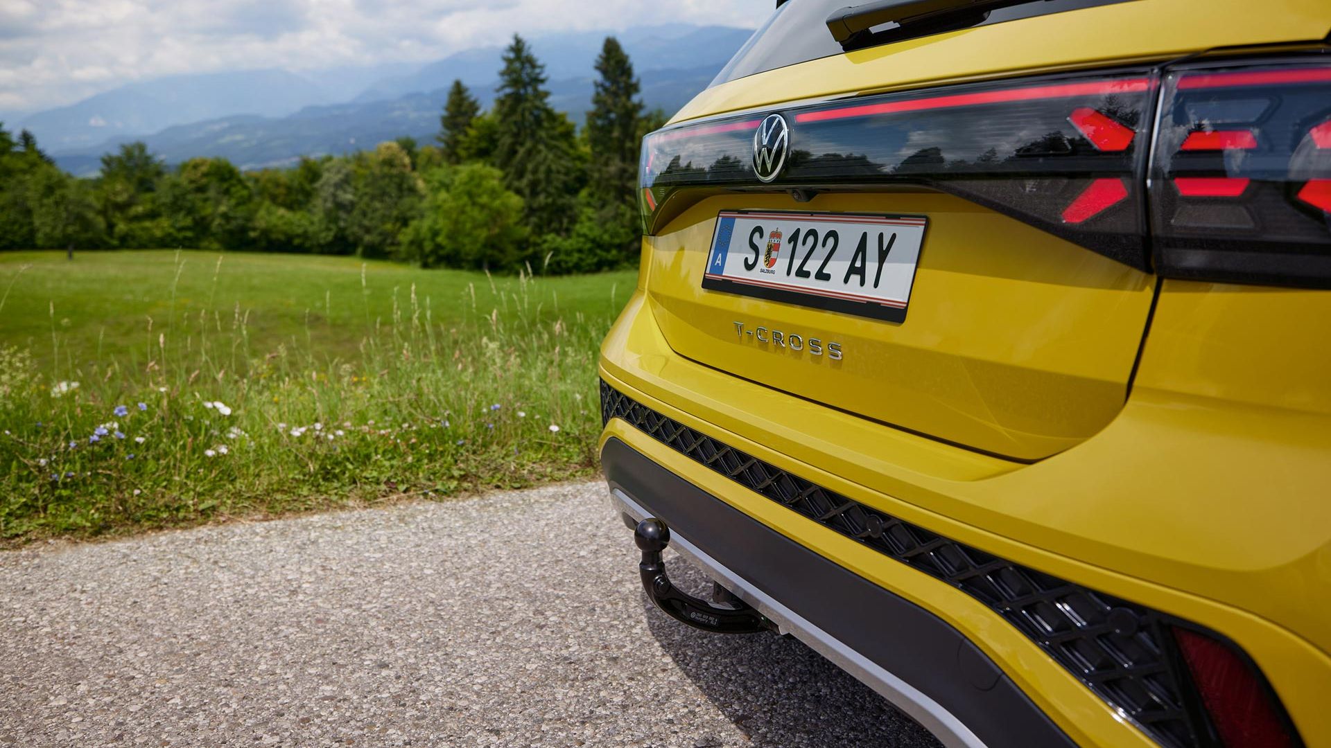 Detail na dvere batožinového priestoru žltého VW T-Cross, ktorý stojí na ceste. V pozadí vidieť lúku, les a hory.