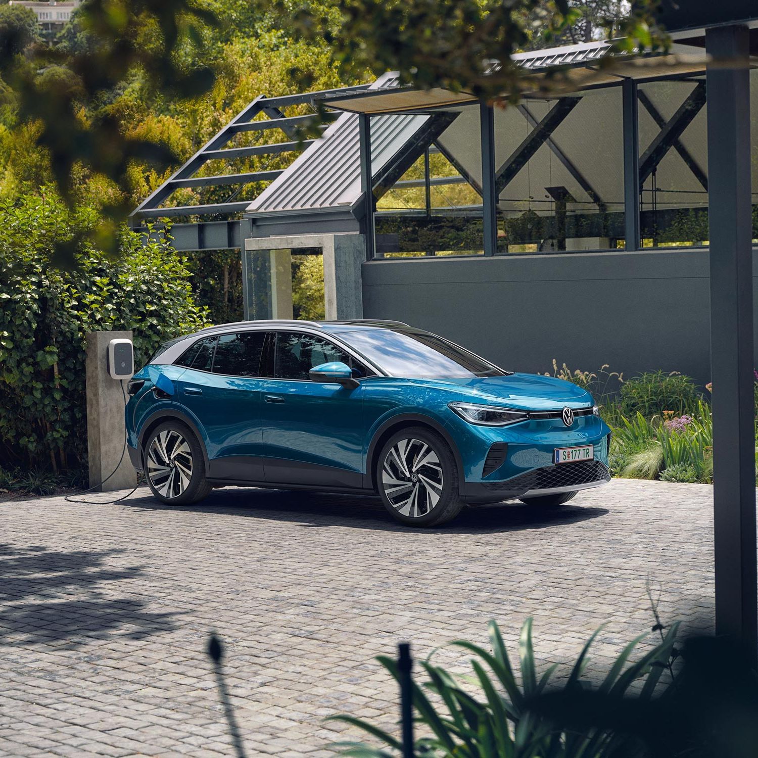 Modré elektrické SUV značky Volkswagen stojí na dláždenej ploche pri domácej nabíjacej stanici. V pozadí je zeleň a malá moderná budova.