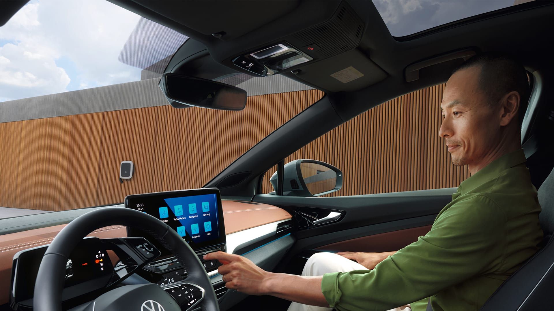 Muž sedí na mieste spolujazdca v elektrickom vozidle značky Volkswagen. Prostredníctvom displeja na palubnej doske ovláda funkcie automobilu. V popredí vidieť pohľad na volant s logom Volkswagen, v pozadí je drevený obklad budovy s nabíjacou stanicou.
