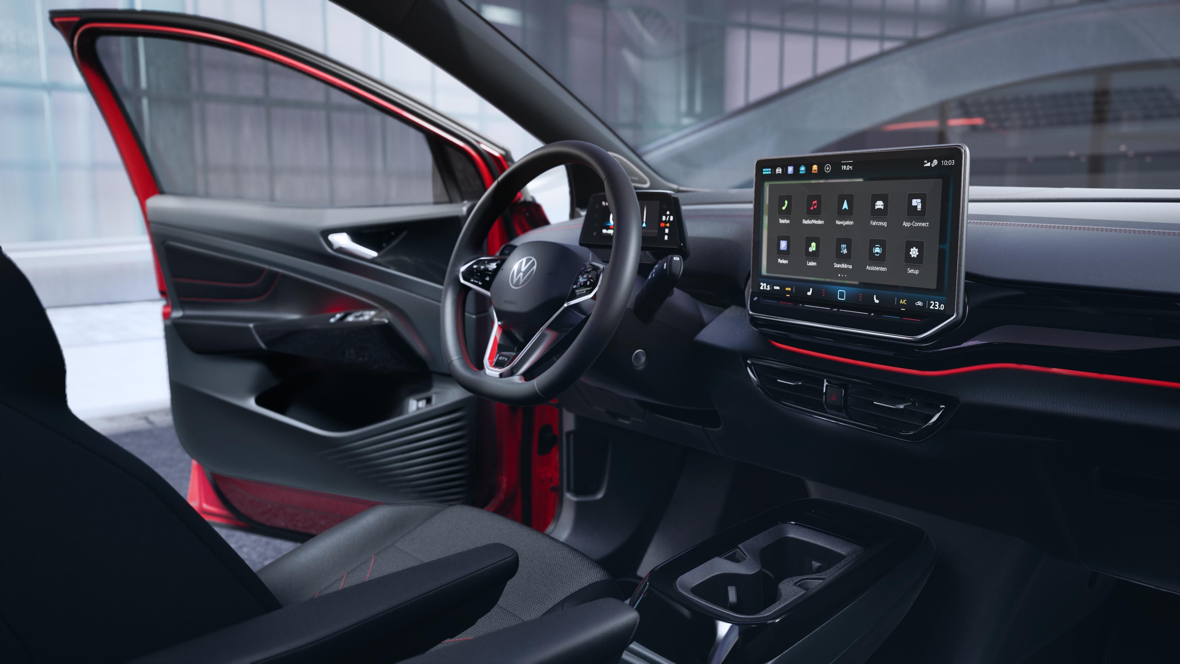 VW ID.4 GTX interiér s výhľadom na predné sedadlá, multifunkčný volant, digitálny prístrojový displej, palubná doska s displejom systému infotainment a ambientné osvetlenie. Dvere vodiča sú otvorené a ponúkajú výhľad na budovu, pri ktorej vozidlo stojí.