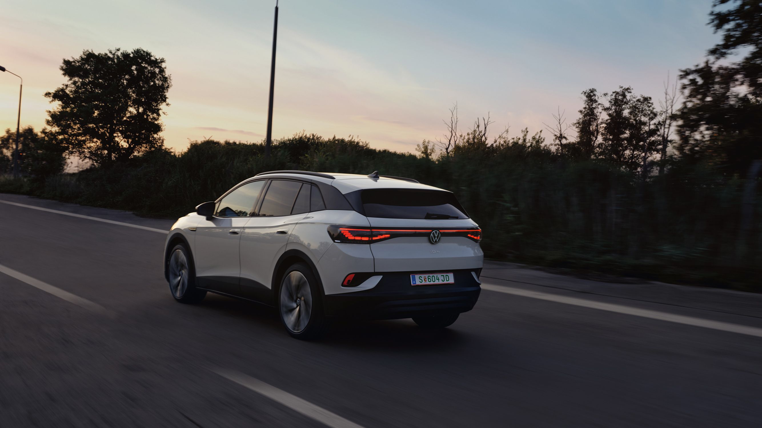 Biely elektrický Volkswagen ID.4 jazdí po ceste za súmraku. Okraj cesty lemujú stromy a kríky. Cesta je rovná a osvetlená pouličnými lampami.