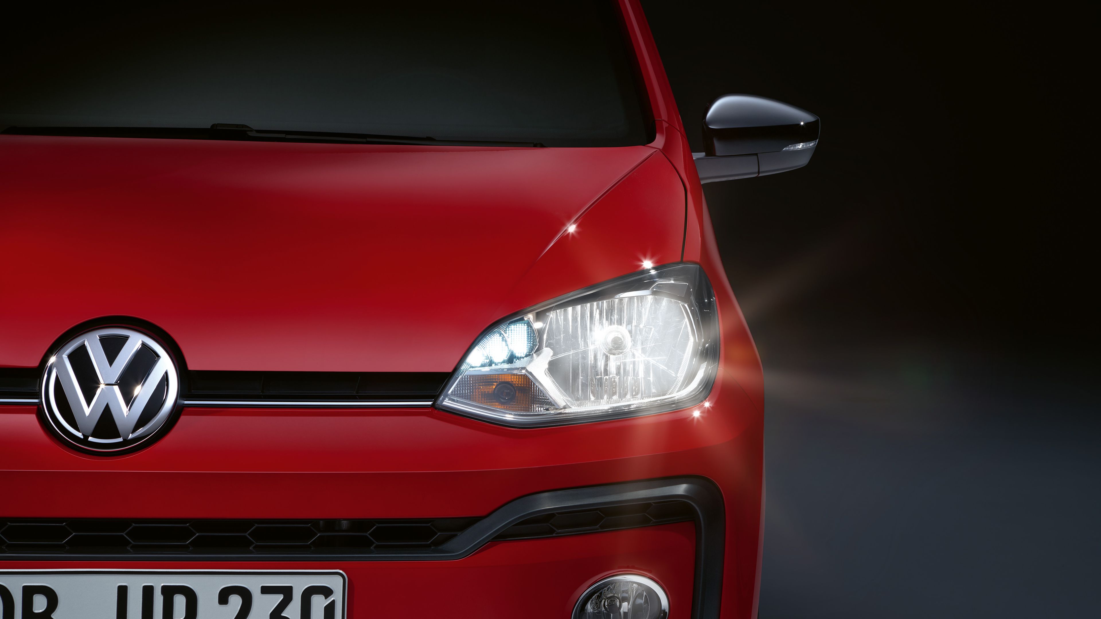 Predná časť červeného vozidla Volkswagen s rozsvietenými prednými svetlami. Viditeľná je registračná značka. Pozadie je tmavé.