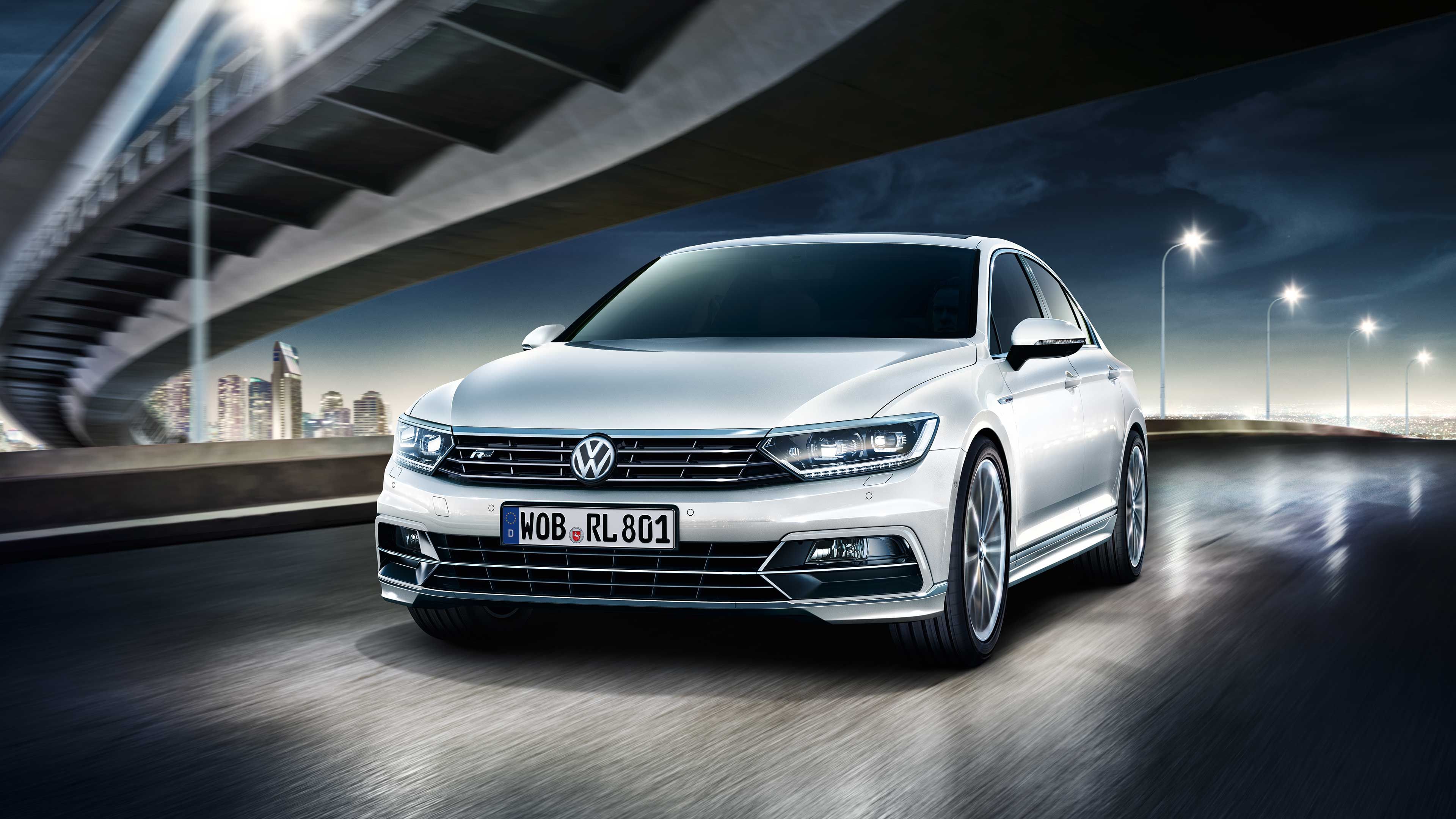 Biele auto značky Volkswagen jazdí po osvetlenej ceste pod mostom. V pozadí je viditeľné centrum mesta s modernými výškovými budovy. Na evidenčnom čísle je nápis "WOB RL801".
