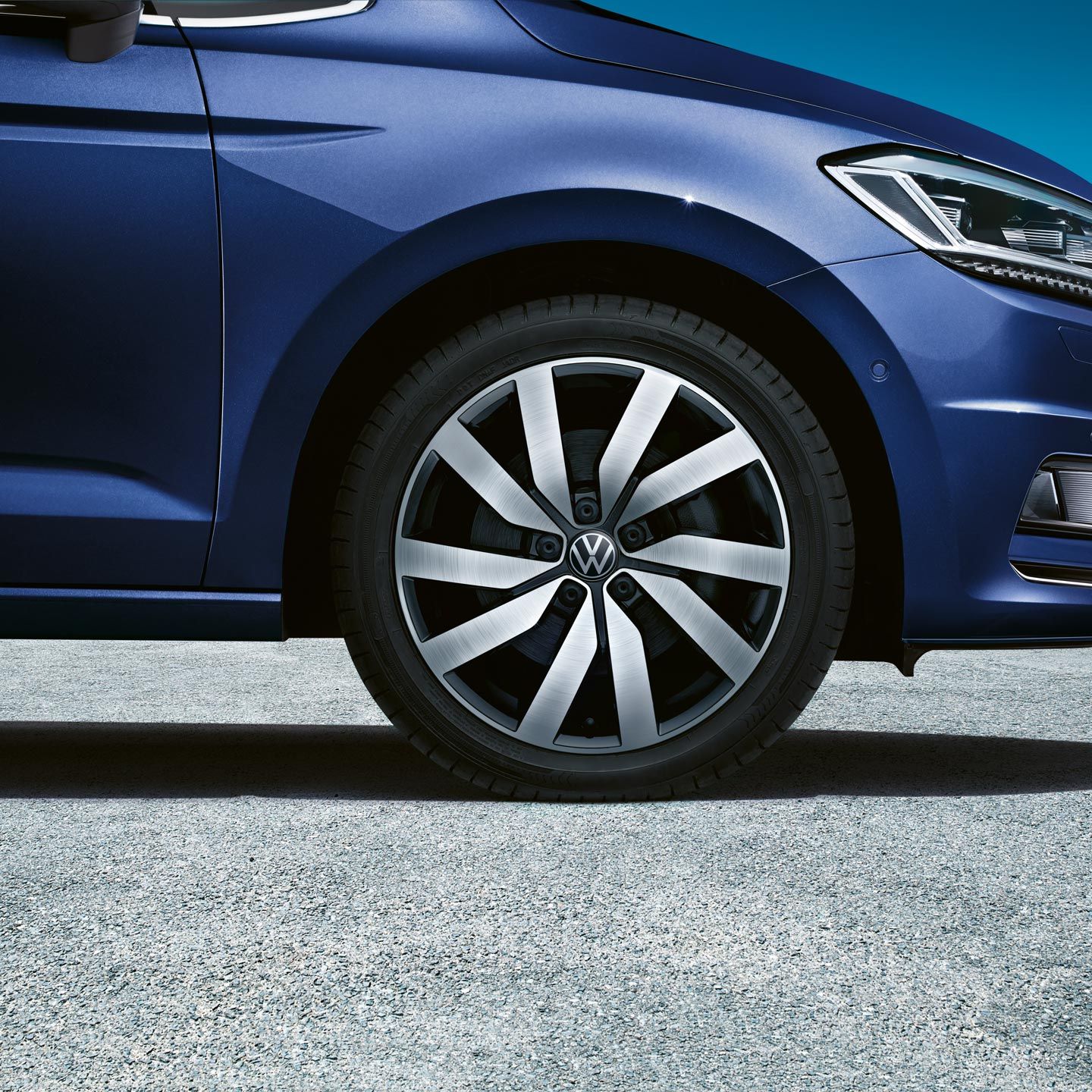 Detailný pohľad na prednú časť modrého vozidla Volkswagen, konkrétne na jeho koleso a disk. Vozidlo stojí na asfaltovej ceste. V pozadí je viditeľná časť krajiny s modrou oblohou.