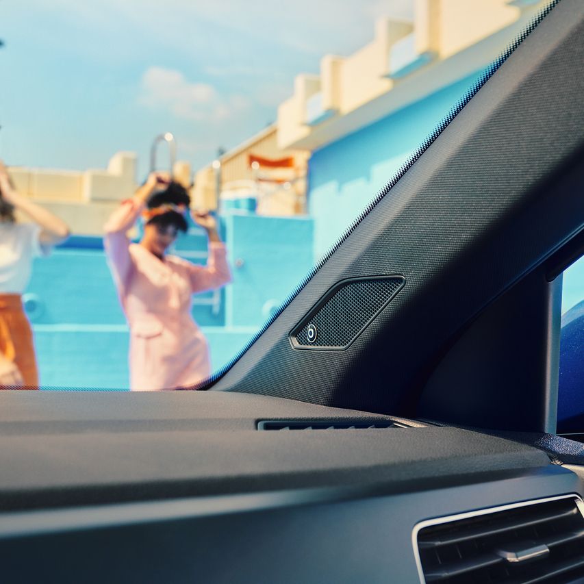 Pohľad do interiéru automobilu na A-stĺpik s integrovaným reproduktorom beats. V pozadí s rozmazaným obrázkom dvoch žien. Okolovidieť svetlomodré steny bazéna.