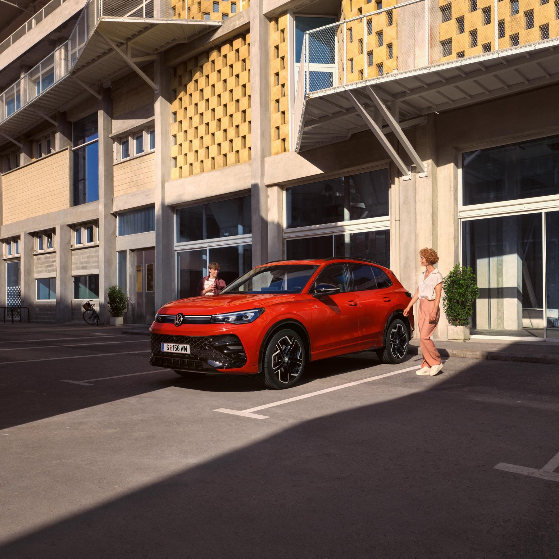 Červený VW Tiguan z bočného pohľadu stojí pred budovou. K vozidlu prichádza žena a chlapec z opačnej strany. Za vozidlom je budova, ktorá vyzerá ako škola.