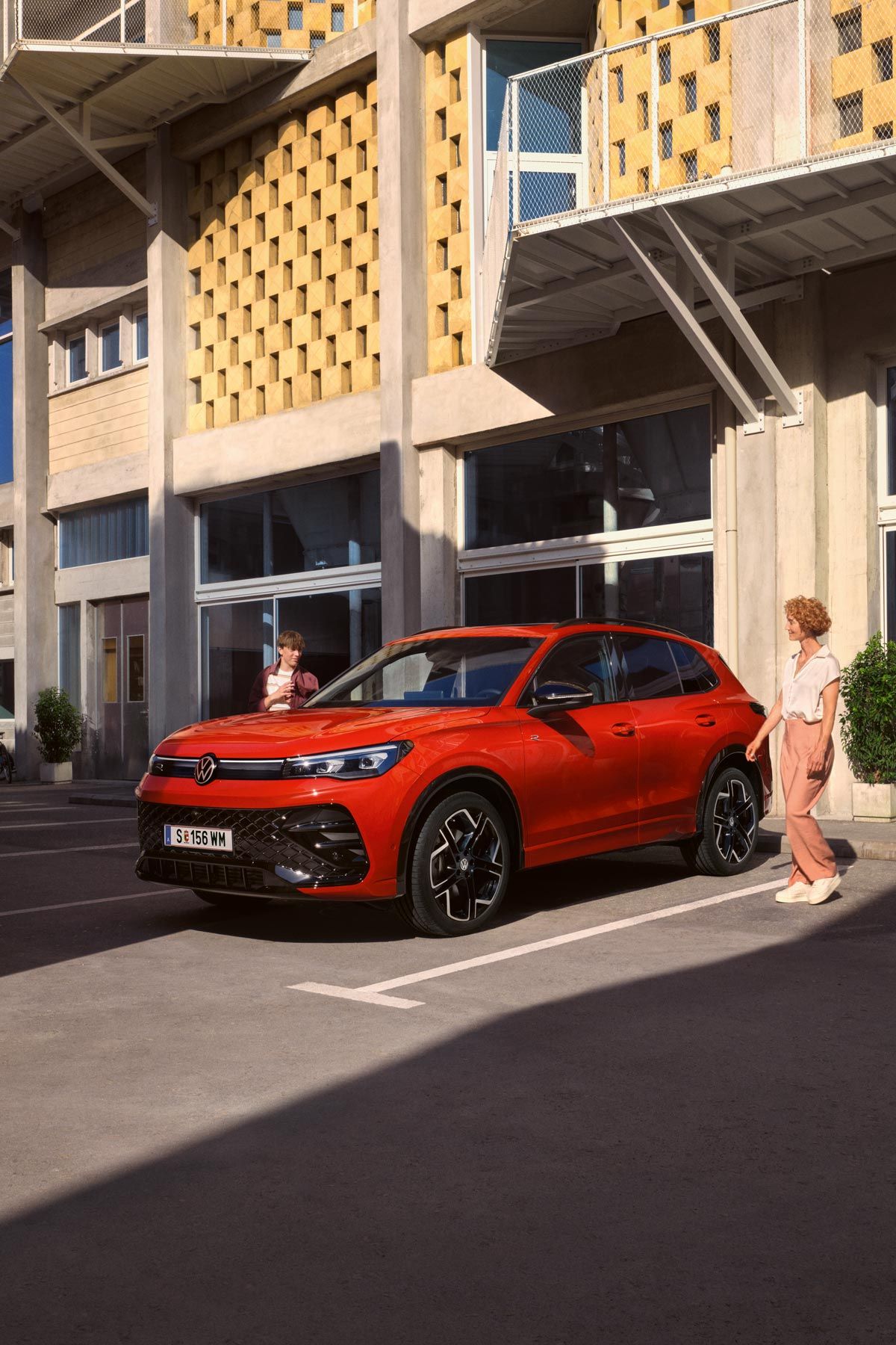 Červený VW Tiguan z bočného pohľadu stojí pred budovou, okolo prechádza žena.