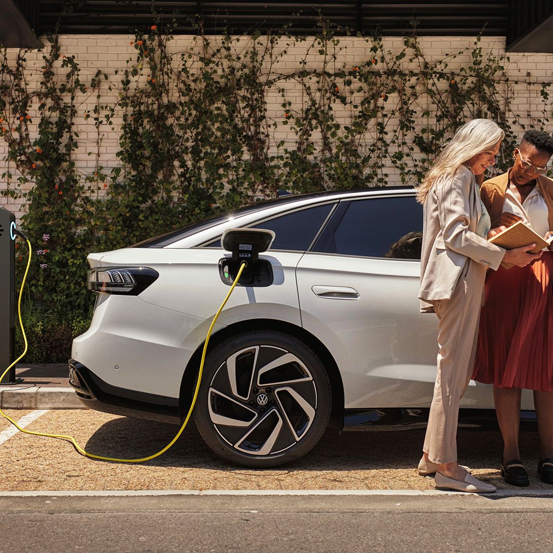 Dve ženy stoja s knihou v ruke vedľa bieleho VW ID., ktorý sa nabíja pri verejnej nabíjacej stanici.