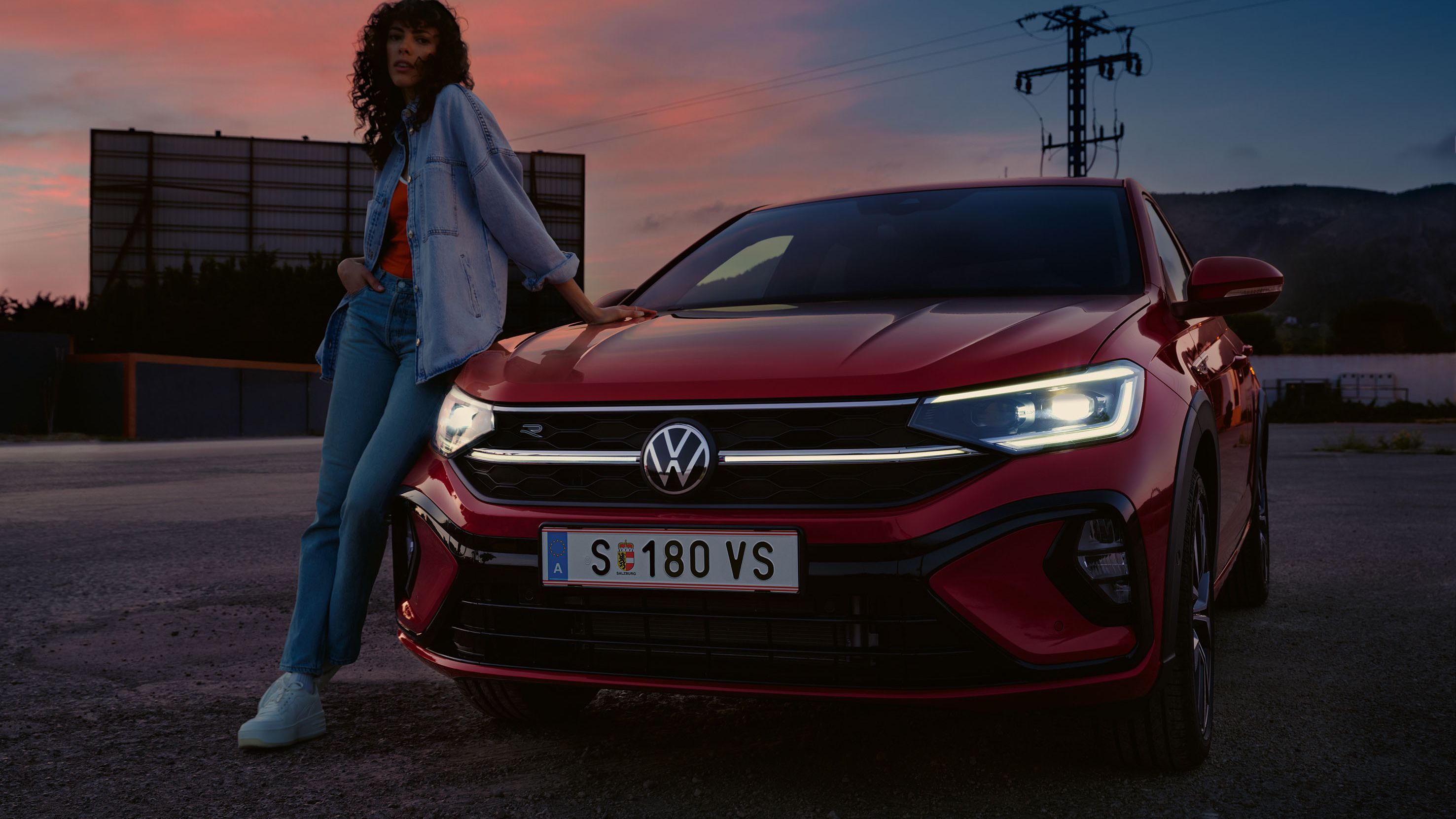 VW Taigo R-Line v červenej farbe na parkovisku. So zapnutými svetlami. Žena opretá o kapotu vozidla. V pozadí vysoká budova, elektrický stĺp a kopce za šera pri západe slnka.