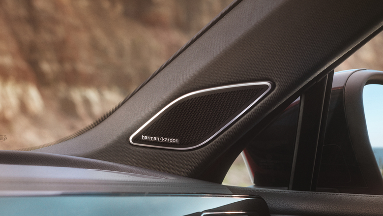 Detailný záber na reproduktor značky Harman Kardon, ktorý je zabudovaný v A-stĺpiku automobilu. Na pozadí je rozmazaný výhľad na vonkajšie prostredie.