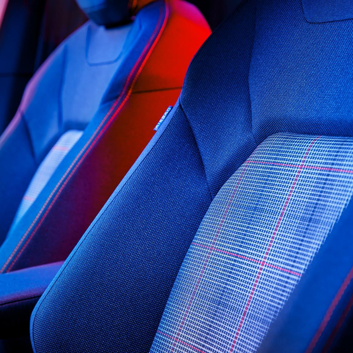 Interiér automobilu so zameraním na predné sedadlá nasvietené  modrým a červeným osvetlením. Sedadlá sú ergonomické s moderným dizajnom a kontrastnými textúrami.