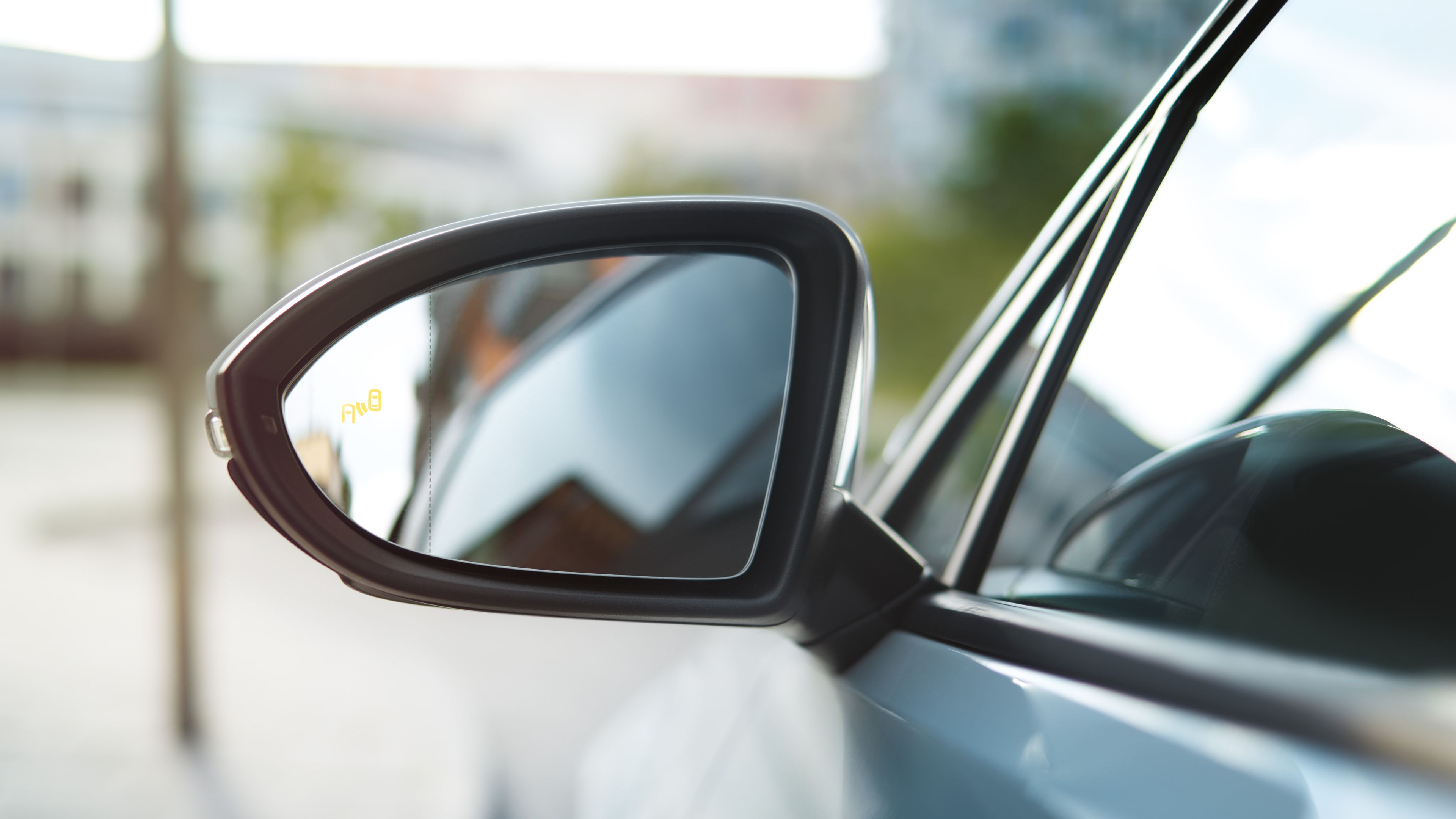 Foto zobrazuje bočné spätné zrkadlo automobilu. Zrkadlo odráža rozmazaný obraz mesta v pozadí. Na zrkadle je viditeľné označenie upozorňujúce na prítomnosť auta v mŕtvom uhle.