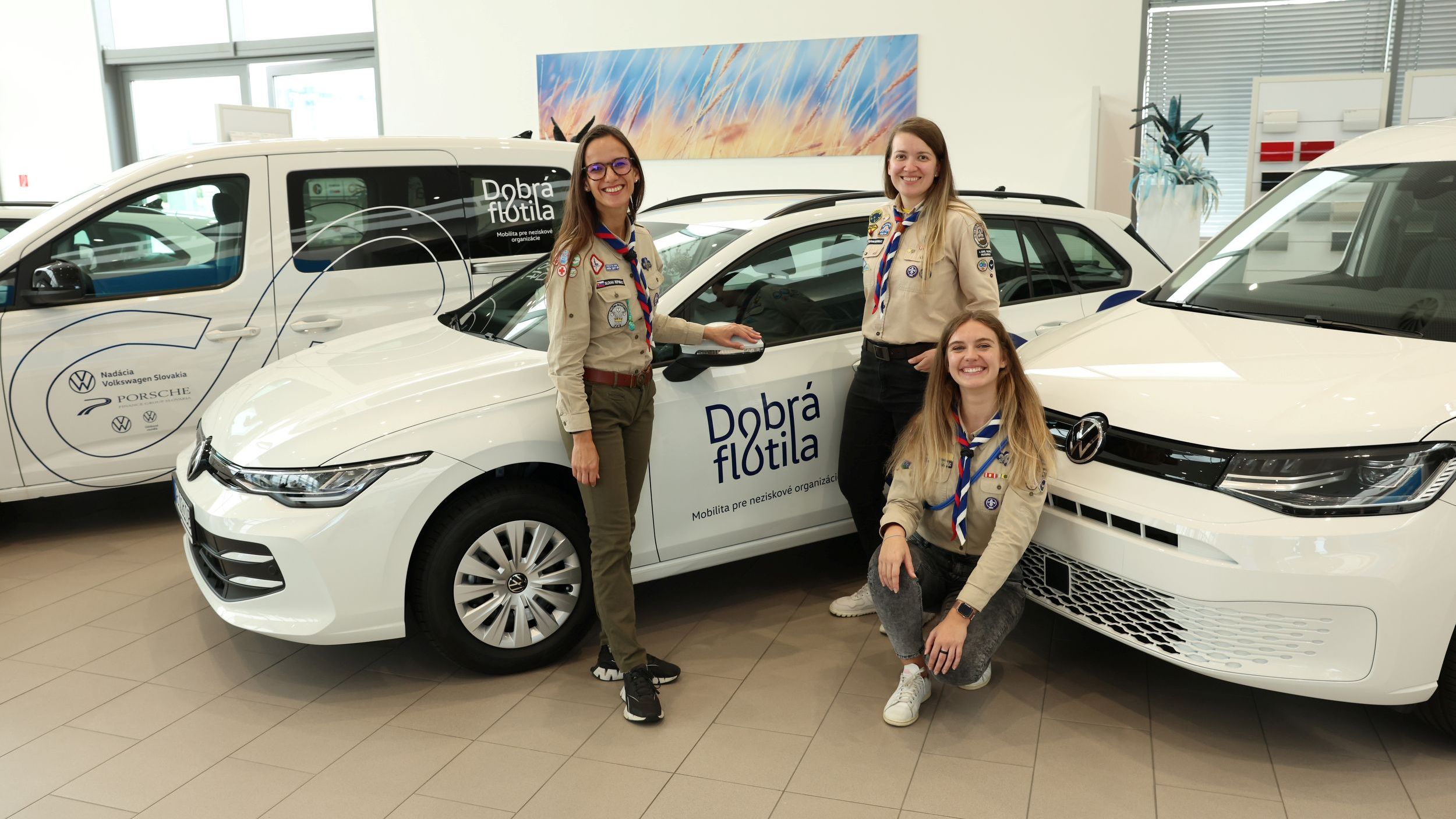 Tri dievčatá v skautských uniformách pózujú pri dvoch automobiloch značky Volkswagen s nápisom "Dobrá flotila" v autosalóne.