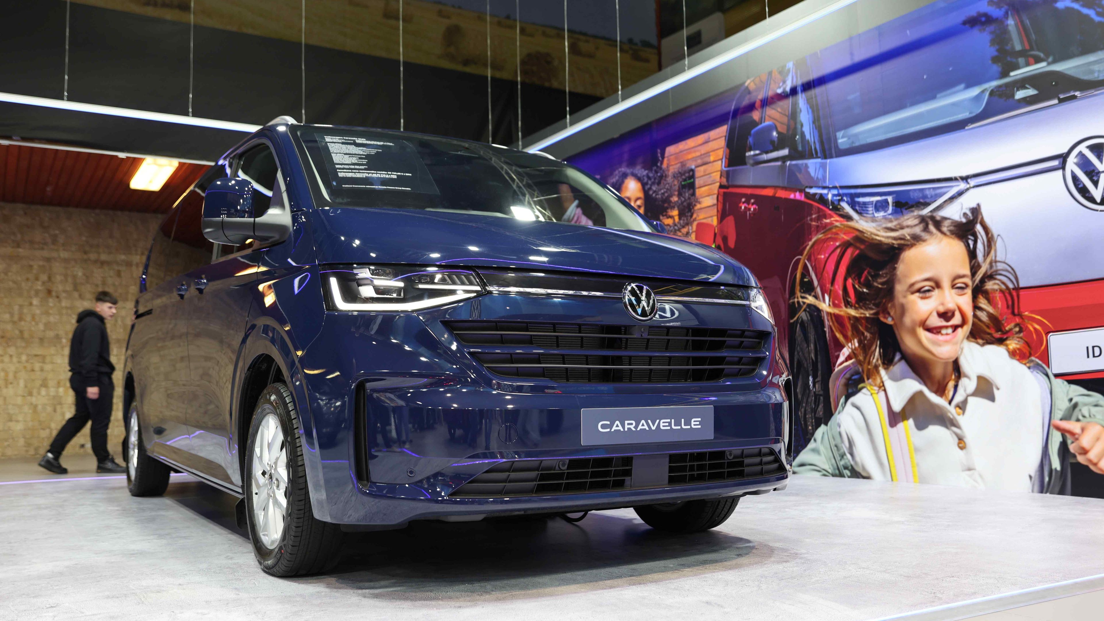 Modré auto Volkswagen Caravelle vystavené na autosalóne v interiéri. Vedľa auta sú reklamné bannery s grafikou a textom. 