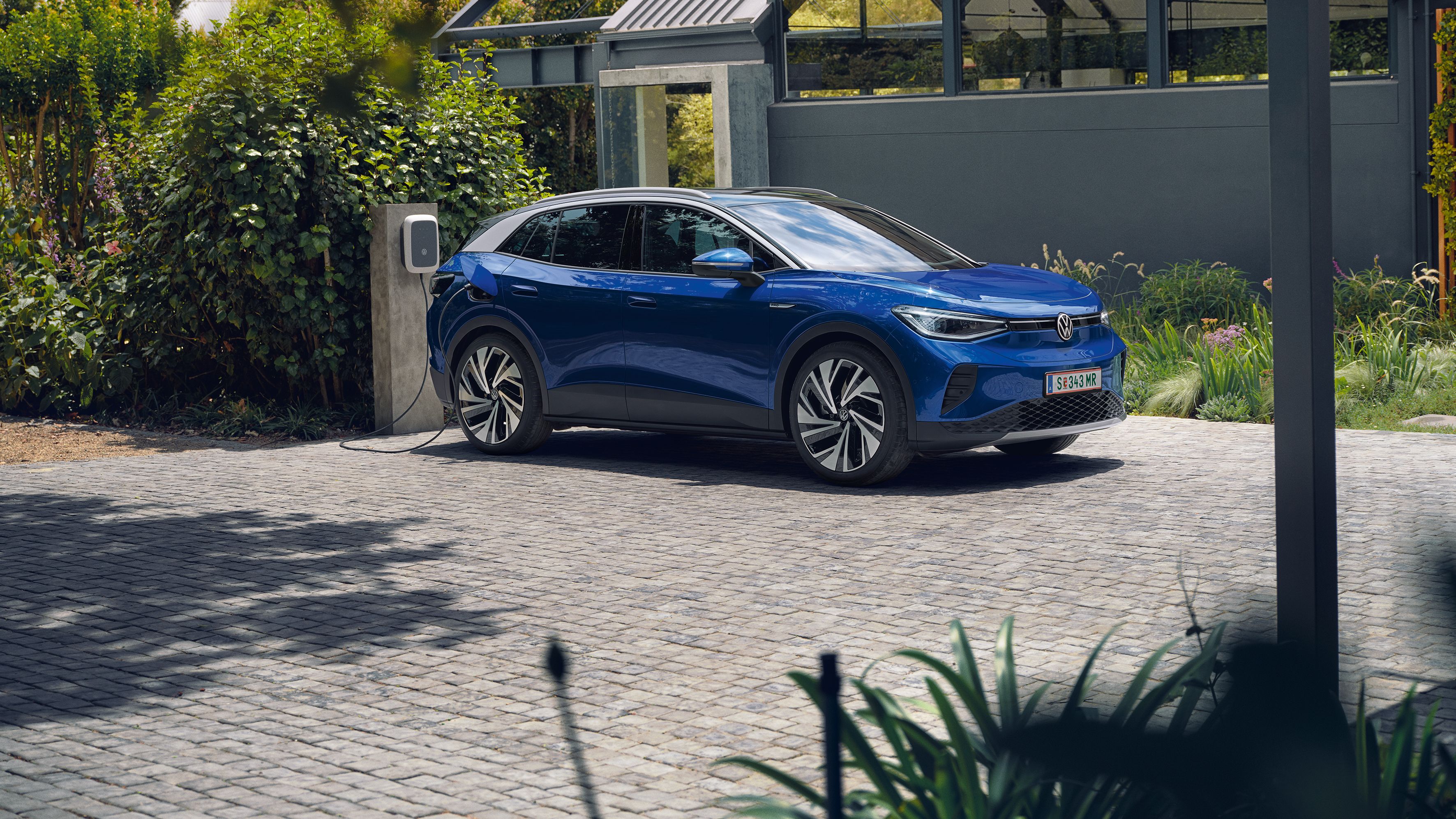 Modré elektrické SUV ID.4 sa nabíja na nabíjacej stanici pred domom. Auto je zaparkované na dláždenom povrchu, okolo ktorého je veľa zelene. V pozadí je moderná stavba.