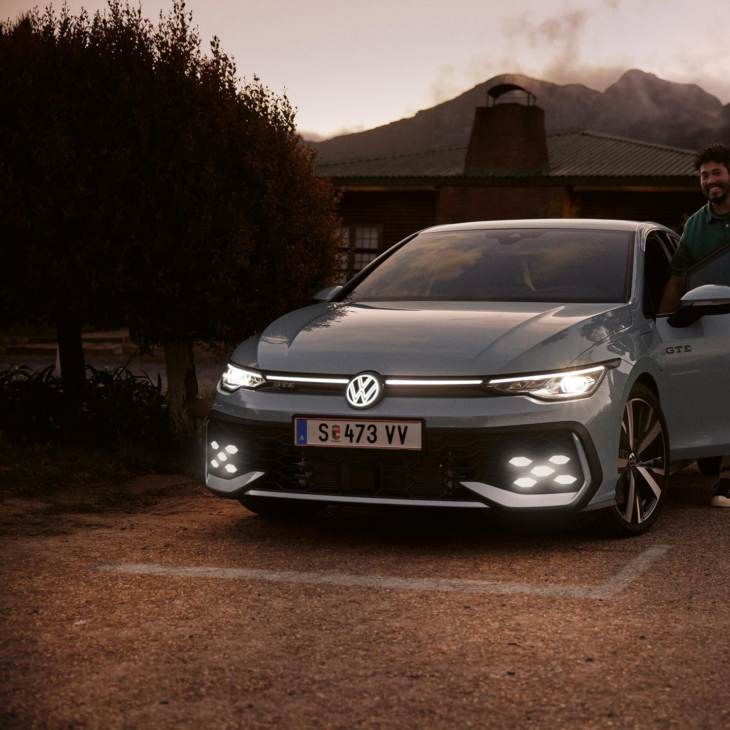 Volkswagen GTE so zasvietenými svetlami stojí na spevnenej ploche pred domom. Vedľa auta stojí muž, ktorý má otvorené dvere vodiča. V pozadí sú viditeľné stromy, dom a hory čiastočne zahalené v hmle.