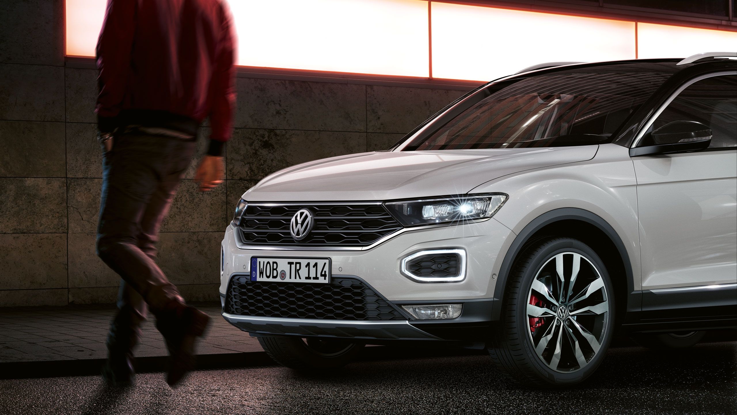 Muž v červenej bunde kráča vedľa bieleho SUV Volkswagen. Auto je zaparkované na mokrej ulici, v pozadí je osvetlená stena.