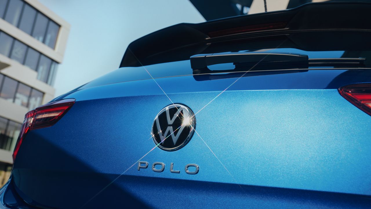 Detailný záber zadnej časti modrého automobilu Volkswagen Polo. Na obrázku sú viditeľné zadné svetlá a logo VW. Pozadie tvorí moderná budova.