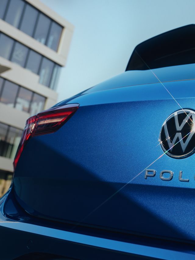 Detailný záber zadnej časti modrého automobilu Volkswagen Polo. Na obrázku sú viditeľné zadné svetlá a logo VW. Pozadie tvorí moderná budova.