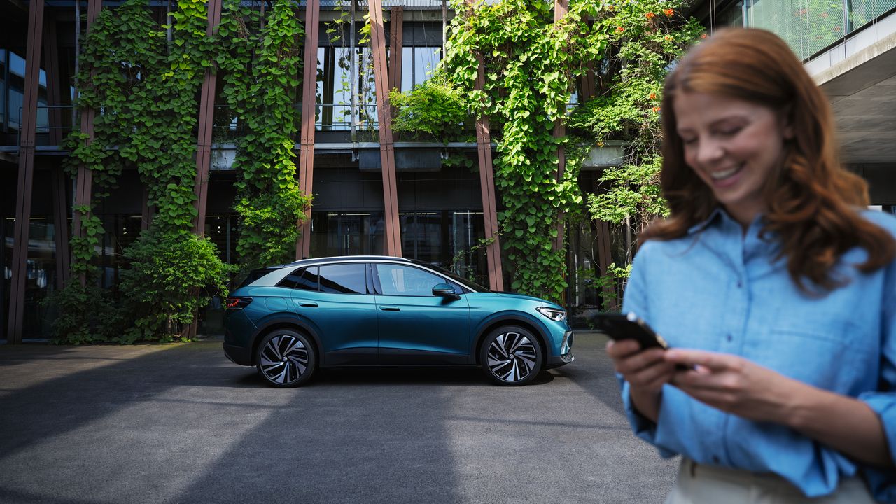 Moderné SUV zaparkované pred budovou pokrytou zeleňou. Žena s rozmazanou tvárou drží telefón a stojí v popredí.